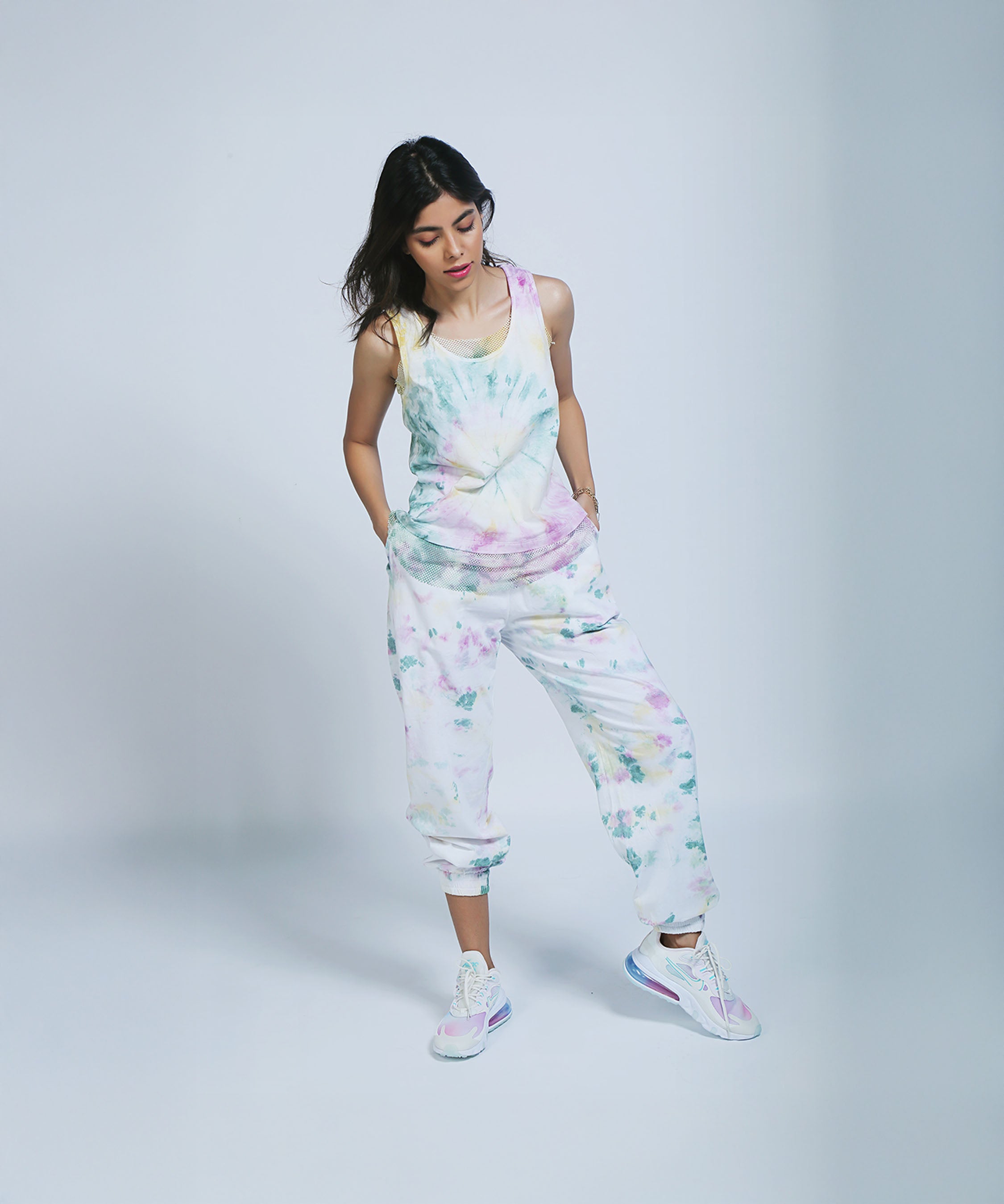 Woman wears Comfort Fit Joggers and a Netted Tank Top, looking down with hands in pockets against a plain background.