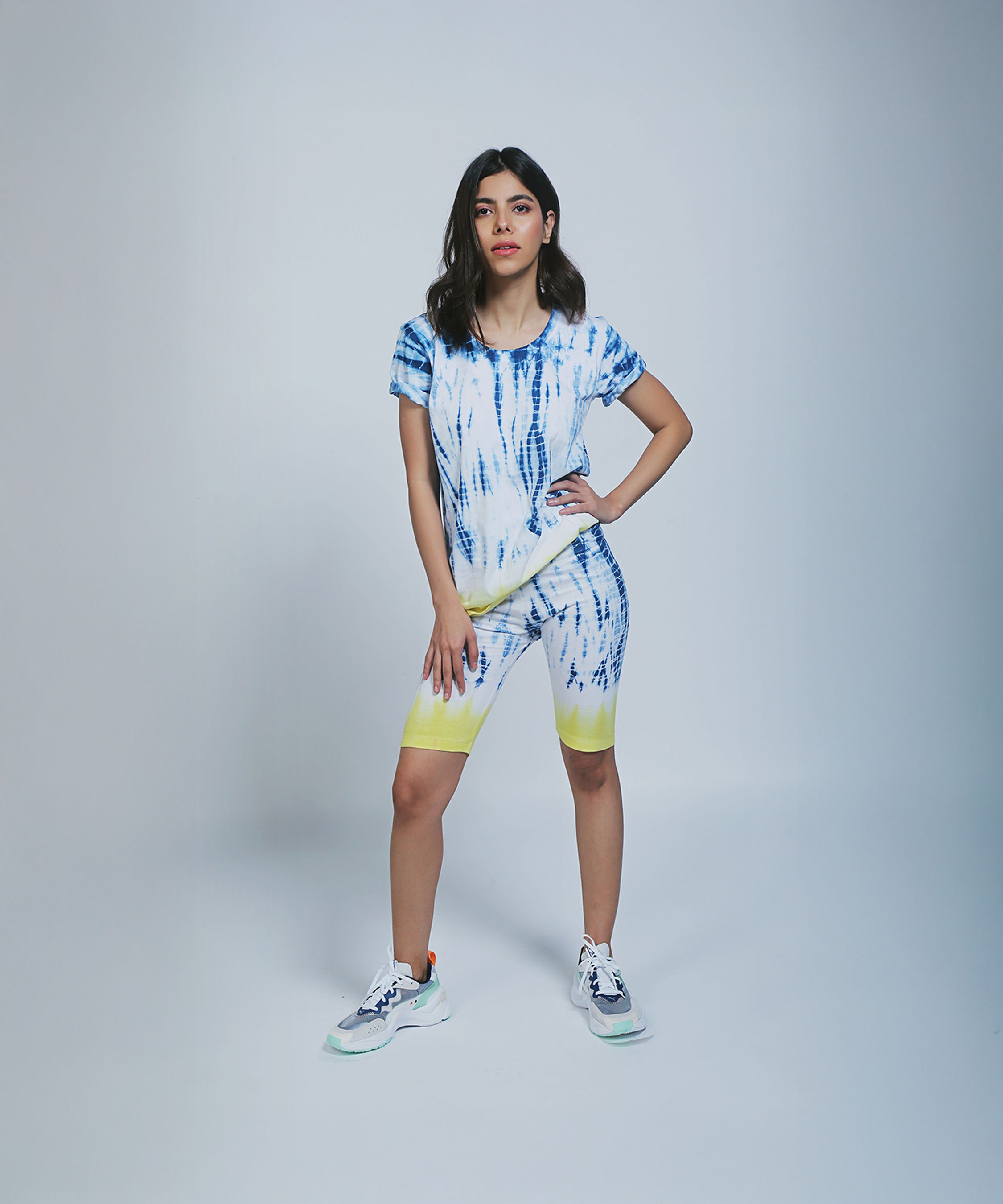 Woman models the Comfort Fit Tee / Sweatshirt + Biker Shorts set, styled with sneakers and one hand on her hip against a plain background.