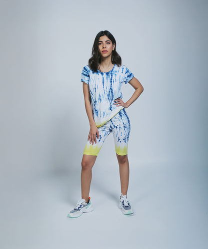 Woman models the Comfort Fit Tee / Sweatshirt + Biker Shorts set, styled with sneakers and one hand on her hip against a plain background.