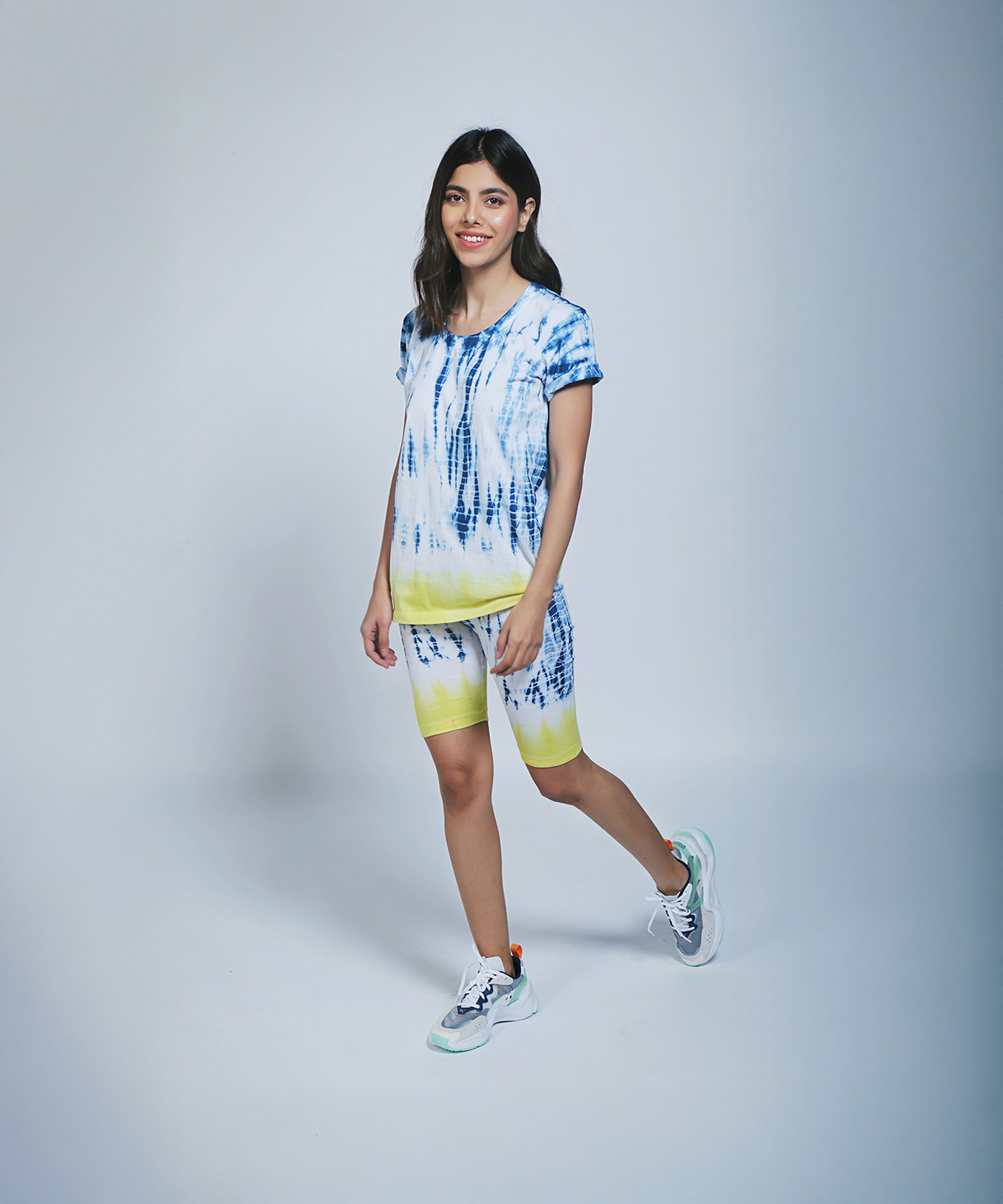 A woman wearing the Comfort Fit Tee / Sweatshirt and Biker Shorts in blue and yellow tie-dye, standing and smiling against a light background.