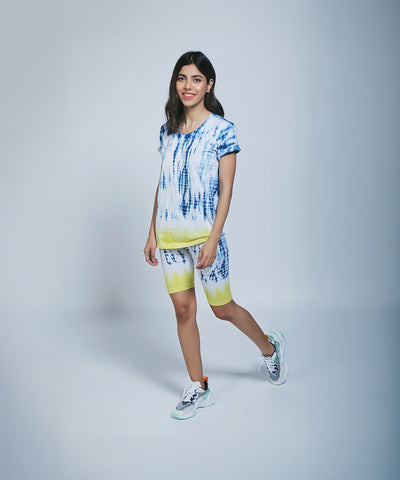 A woman wearing the Comfort Fit Tee / Sweatshirt and Biker Shorts in blue and yellow tie-dye, standing and smiling against a light background.