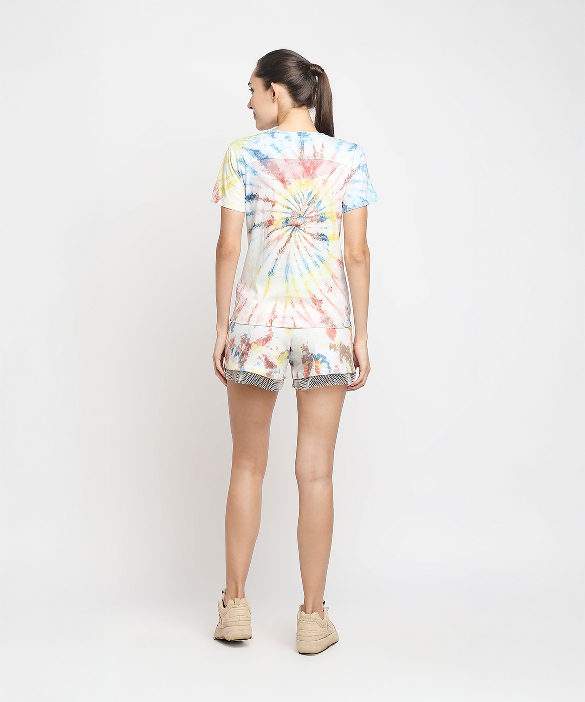 A woman stands with her back to the camera, wearing the Netted shorts + comfort tee set featuring high-waisted netted shorts and a vibrant tie-dye cotton comfort tee, paired with beige sneakers.