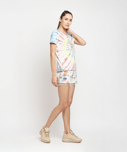 Woman poses against a plain white background wearing the Netted shorts + comfort tee in colorful tie-dye, paired with beige sneakers. Eco-friendly dyes give her cotton outfit vibrant flair.