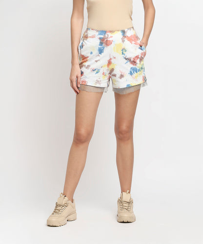A woman wears Netted Shorts with a colorful abstract print, made using eco-friendly dyes, paired with the Comfort Tee in beige and matching sneakers, hands in pockets.