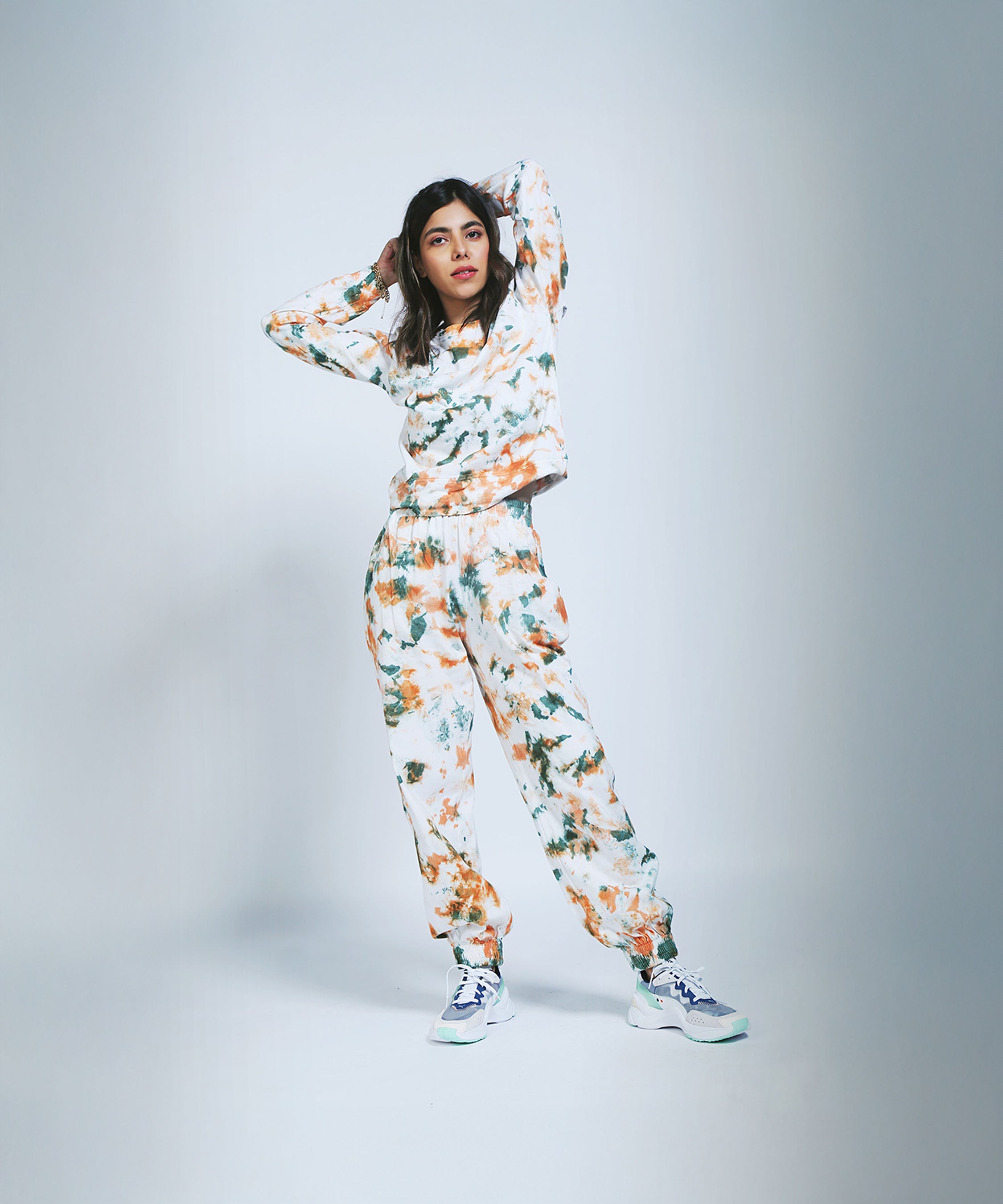 A woman in an Earthy Tones Sweatshirt / Tee and Comfort Joggers with white sneakers stands hands behind her head against a light backdrop.