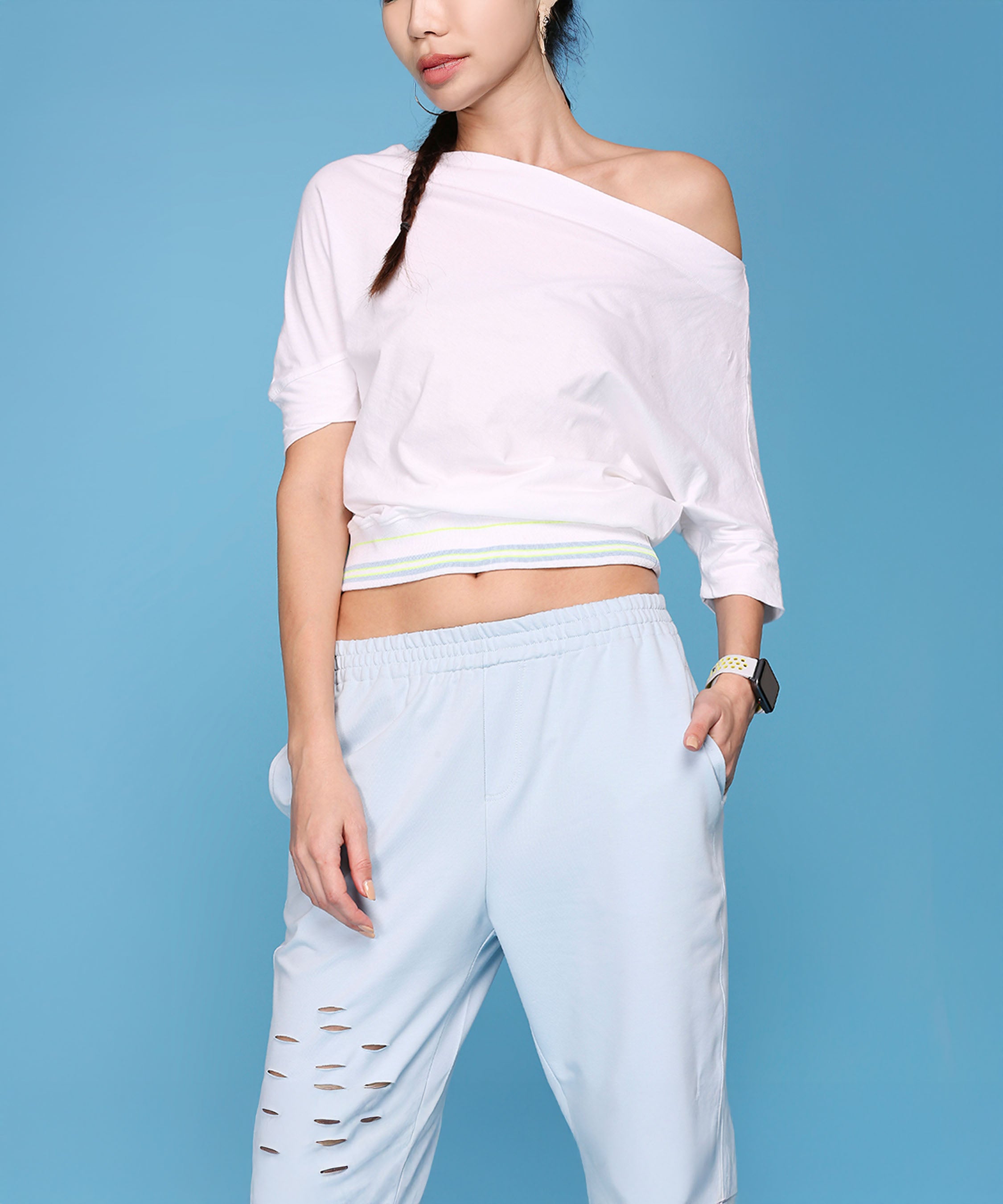 A woman stands against a blue background wearing the Off Shoulder White Rib/Light Blue Ripped Joggers AW21.