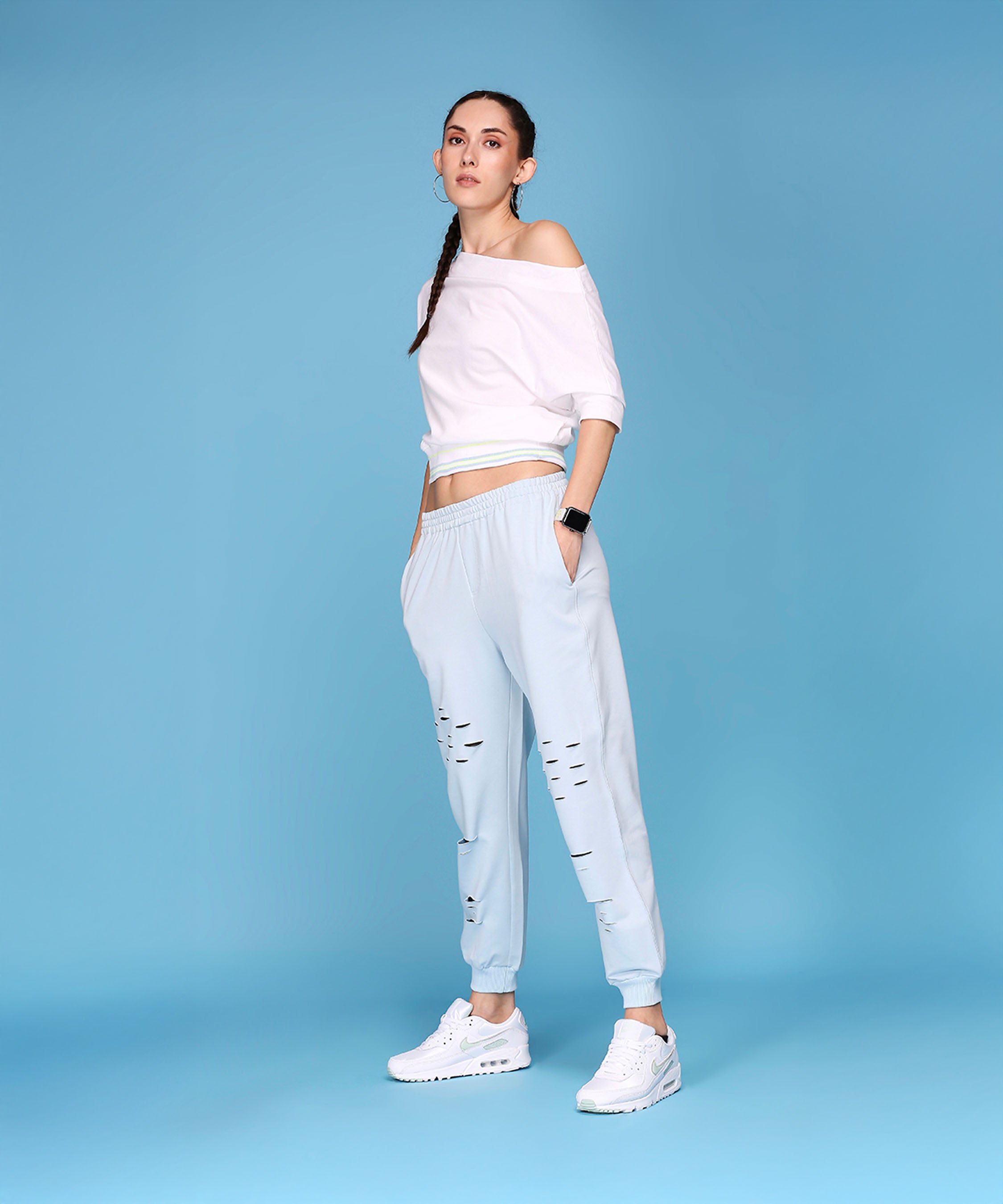 A woman wears the Off Shoulder White Rib/Light Blue Ripped Joggers AW21, standing against a solid light blue background, highlighting these comfy joggers for a relaxed yet stylish look.