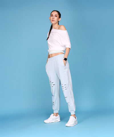 A woman wears the Off Shoulder White Rib/Light Blue Ripped Joggers AW21, standing against a solid light blue background, highlighting these comfy joggers for a relaxed yet stylish look.