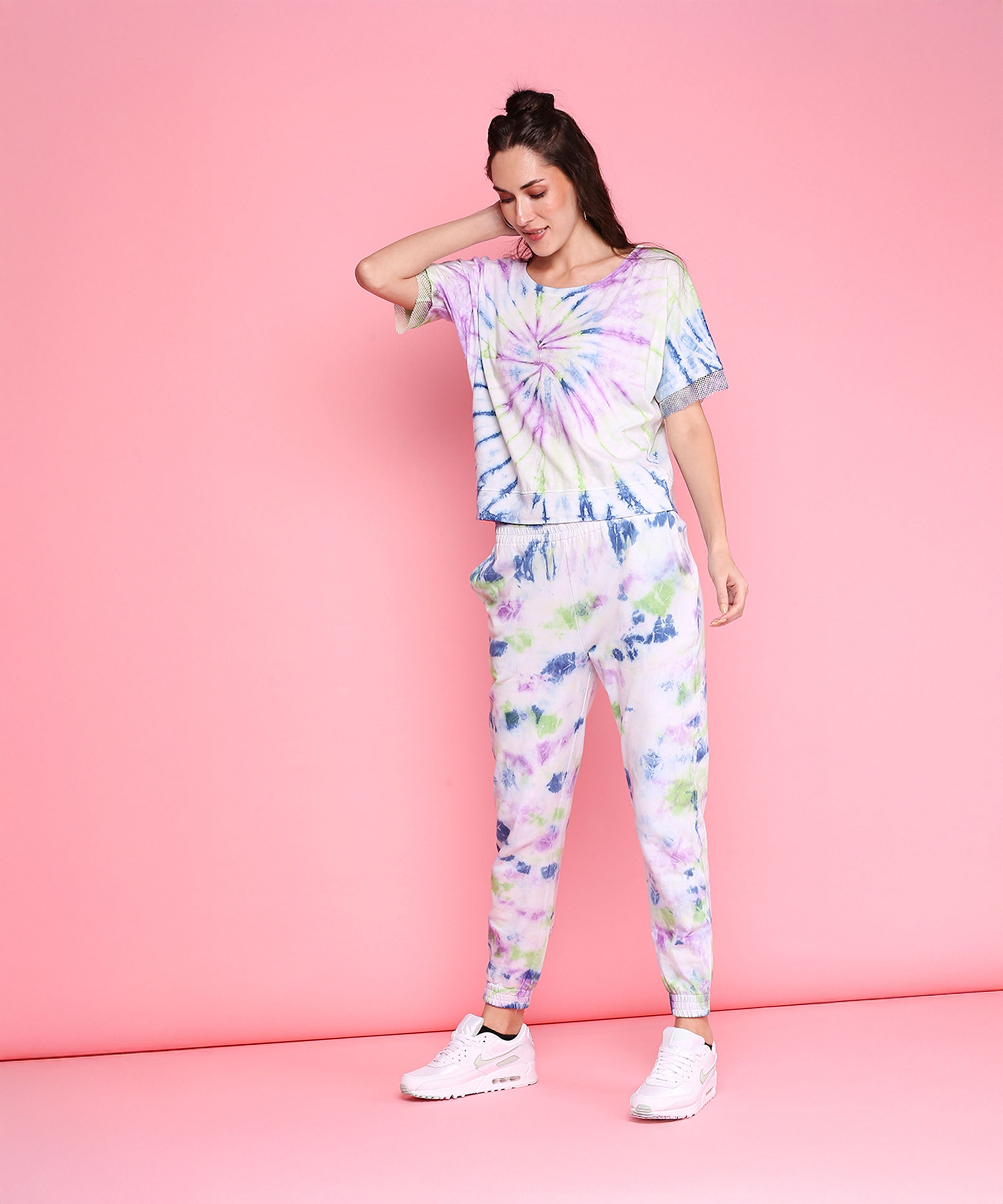 A woman models the Comfort Fit Tee + Joggers - AW21, a breathable, hand-dyed pastel tie-dye set with an oversized tee and joggers, paired with white sneakers against a solid pink background.