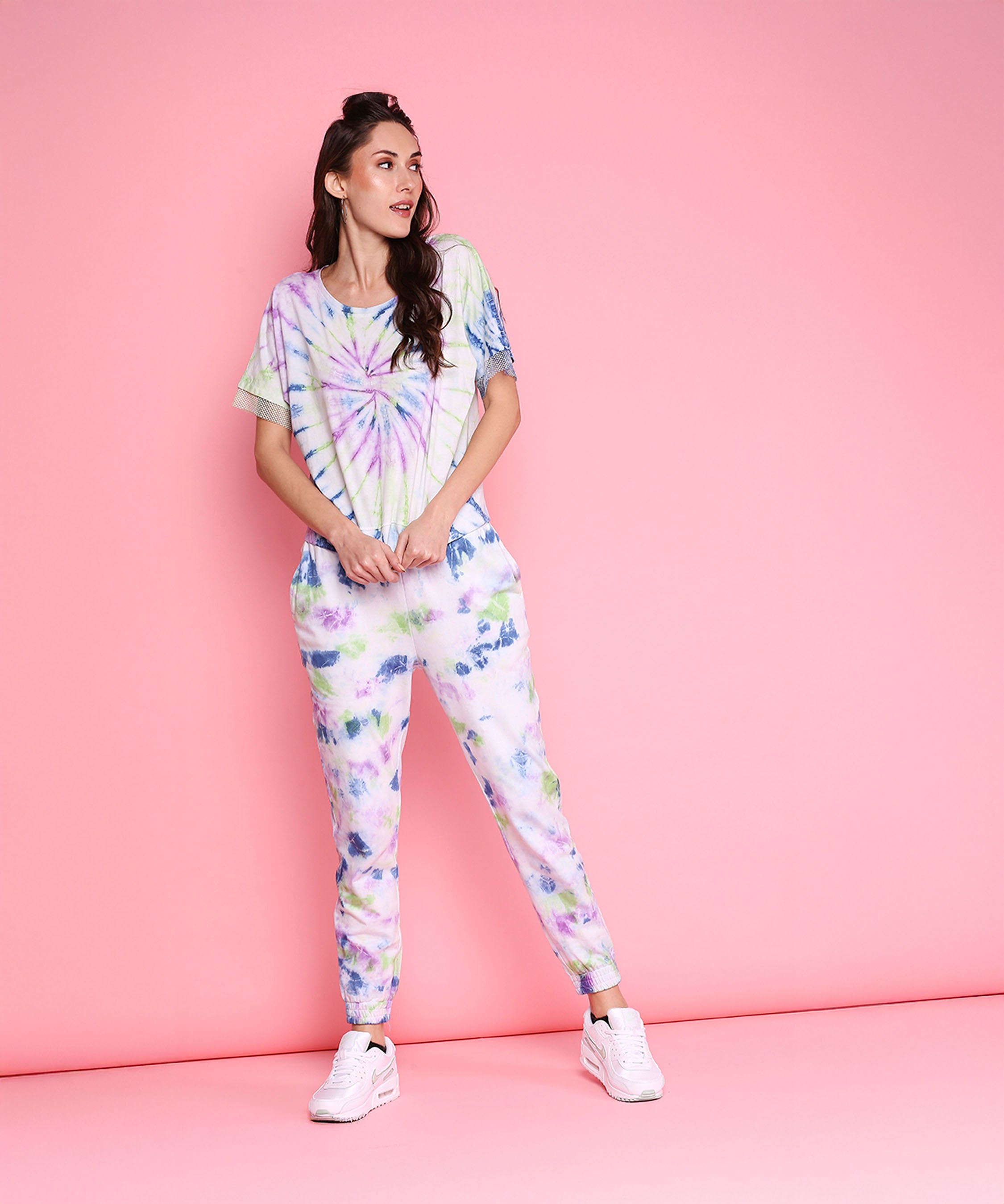 A woman wears the Comfort Fit Tee + Joggers - AW21 with white sneakers, standing against a pink background and looking to the side.