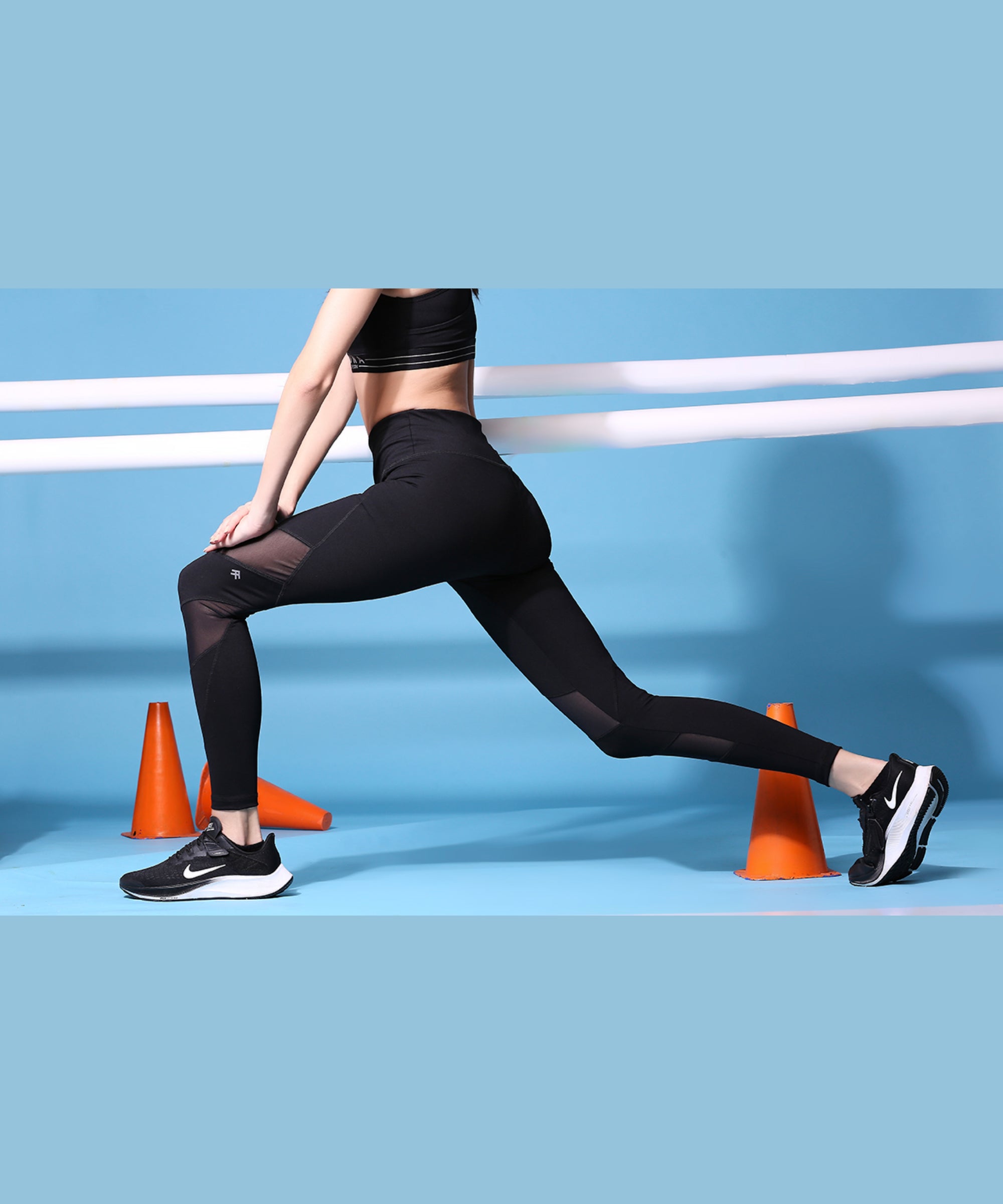 A woman in Netted High Rise Compression Leggings- AW21 performs a lunge stretch near orange cones against a blue background.