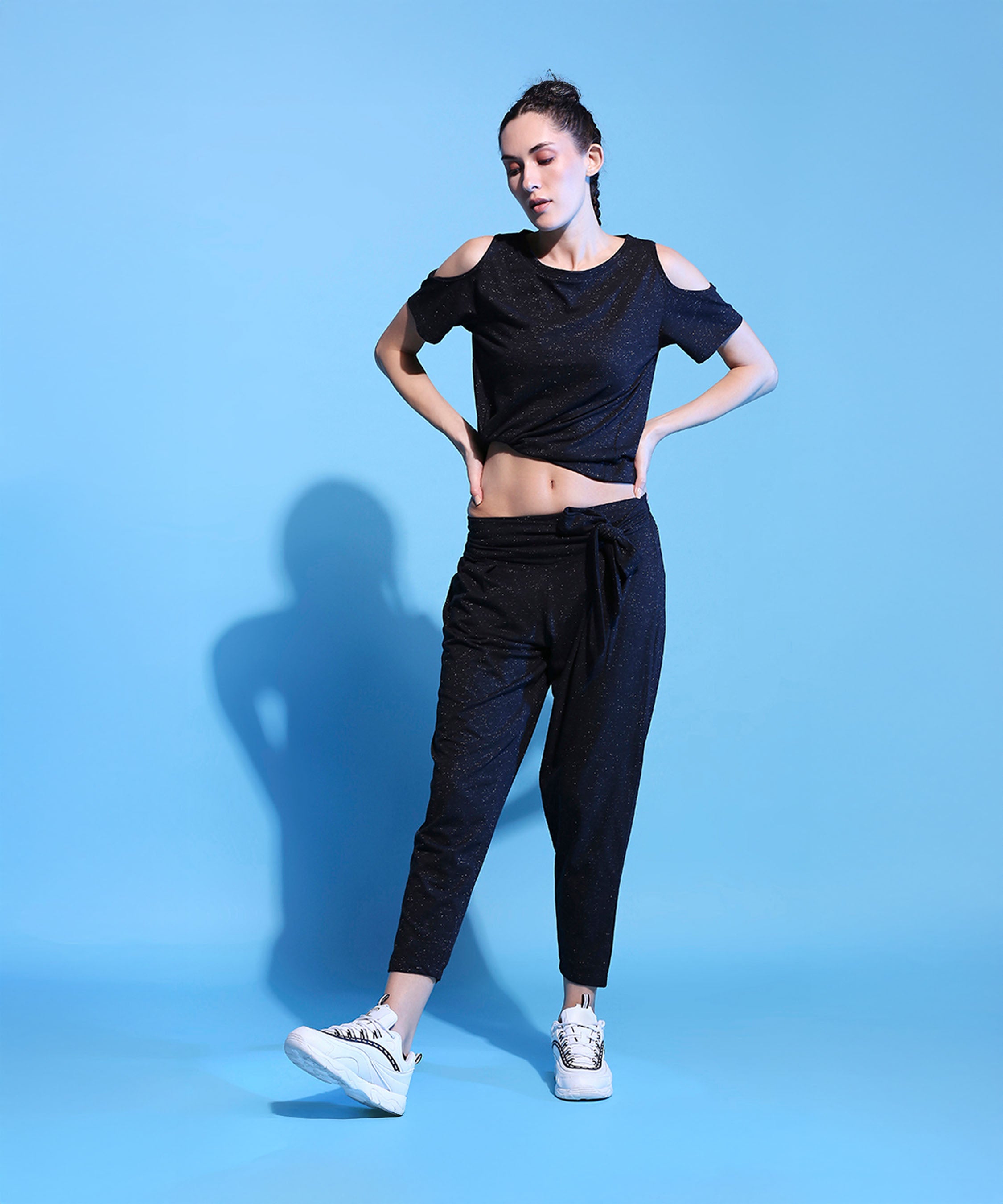 Woman wearing the Shimmer Cold Shoulder Tee (Customised) and Side Tie Up Shimmer Yoga Pants- AW21 with white sneakers, posing against a solid light blue background.