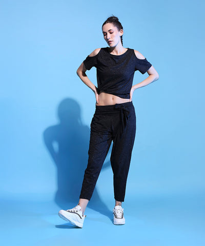 Woman wearing the Shimmer Cold Shoulder Tee (Customised) and Side Tie Up Shimmer Yoga Pants- AW21 with white sneakers, posing against a solid light blue background.