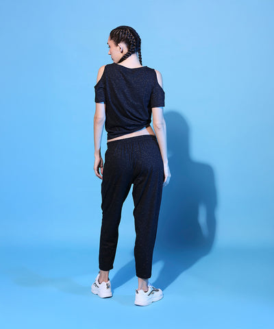 Woman with braided hair wears the Shimmer Cold Shoulder Tee (Customised) and Side Tie Up Shimmer Yoga Pants - AW21, standing on a blue background, facing away.