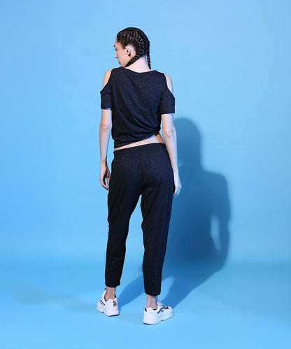 Woman with braided hair wears the Shimmer Cold Shoulder Tee (Customised) and Side Tie Up Shimmer Yoga Pants - AW21, standing on a blue background, facing away.