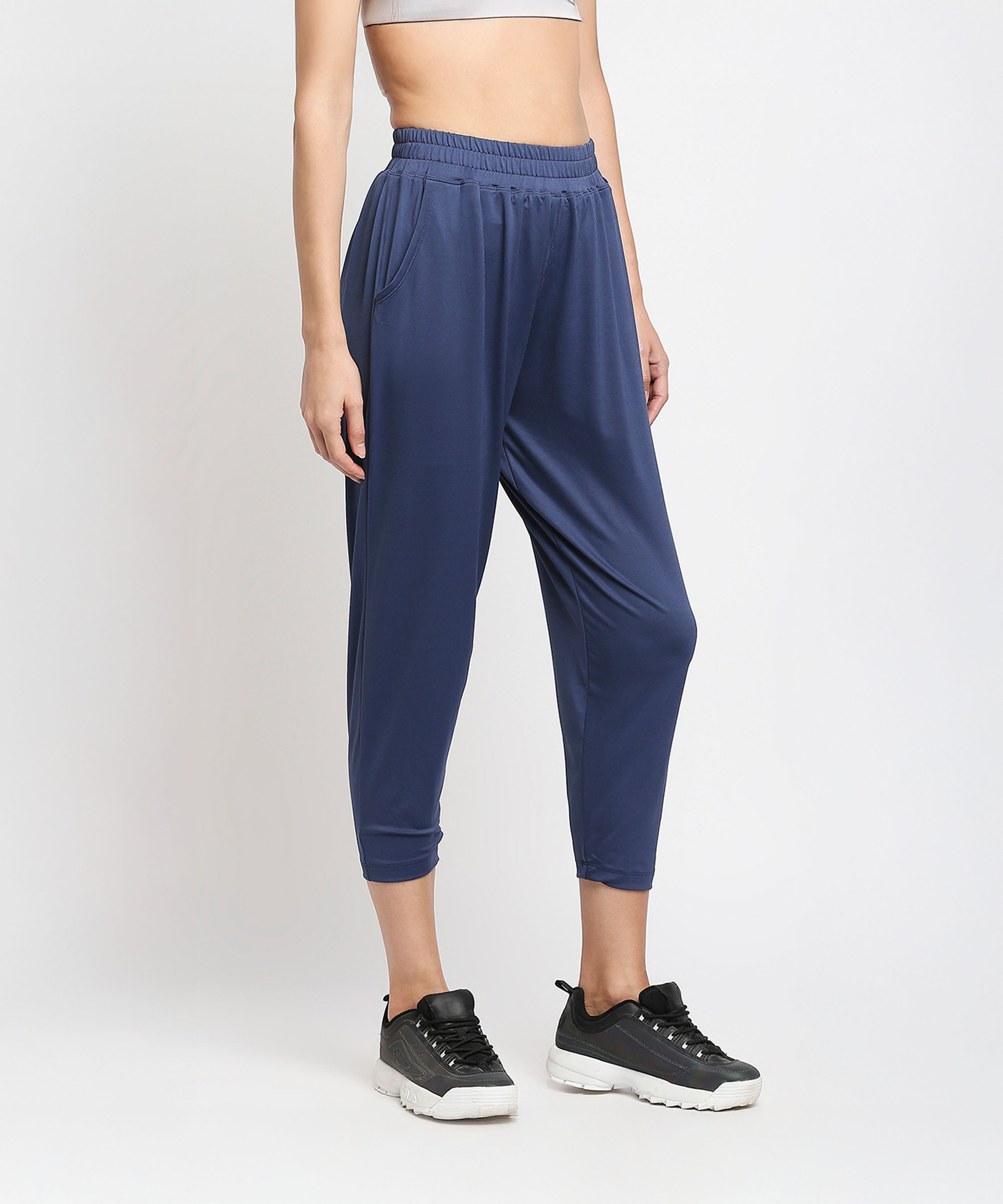 A woman stands against a white background wearing Blue Yoga Pants - AW21, a grey crop top, and black sneakers.