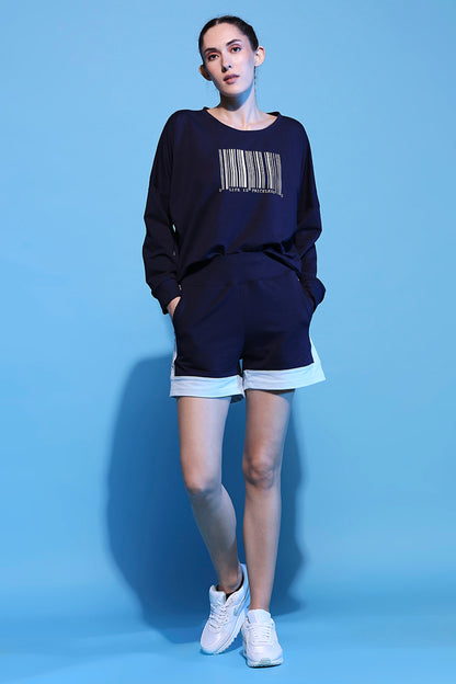 Woman wearing Barcode Printed Sweatshirt + Shorts - AW21 set, featuring a navy sweatshirt with barcode design, matching navy shorts with white trim, and white sneakers, standing on a blue background.