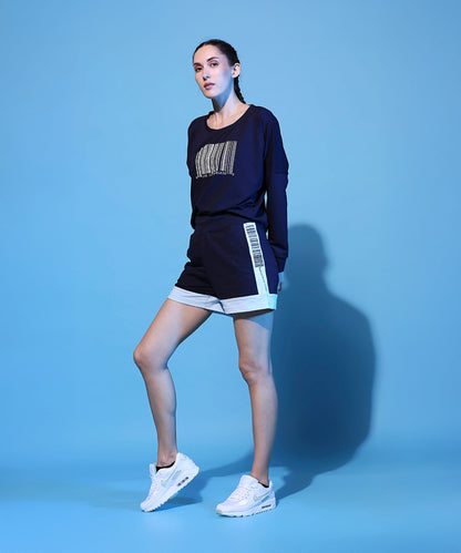 A woman poses confidently against a solid light blue background, wearing a Barcode Printed Navy Blue Sweatshirt, matching shorts, and white sneakers.