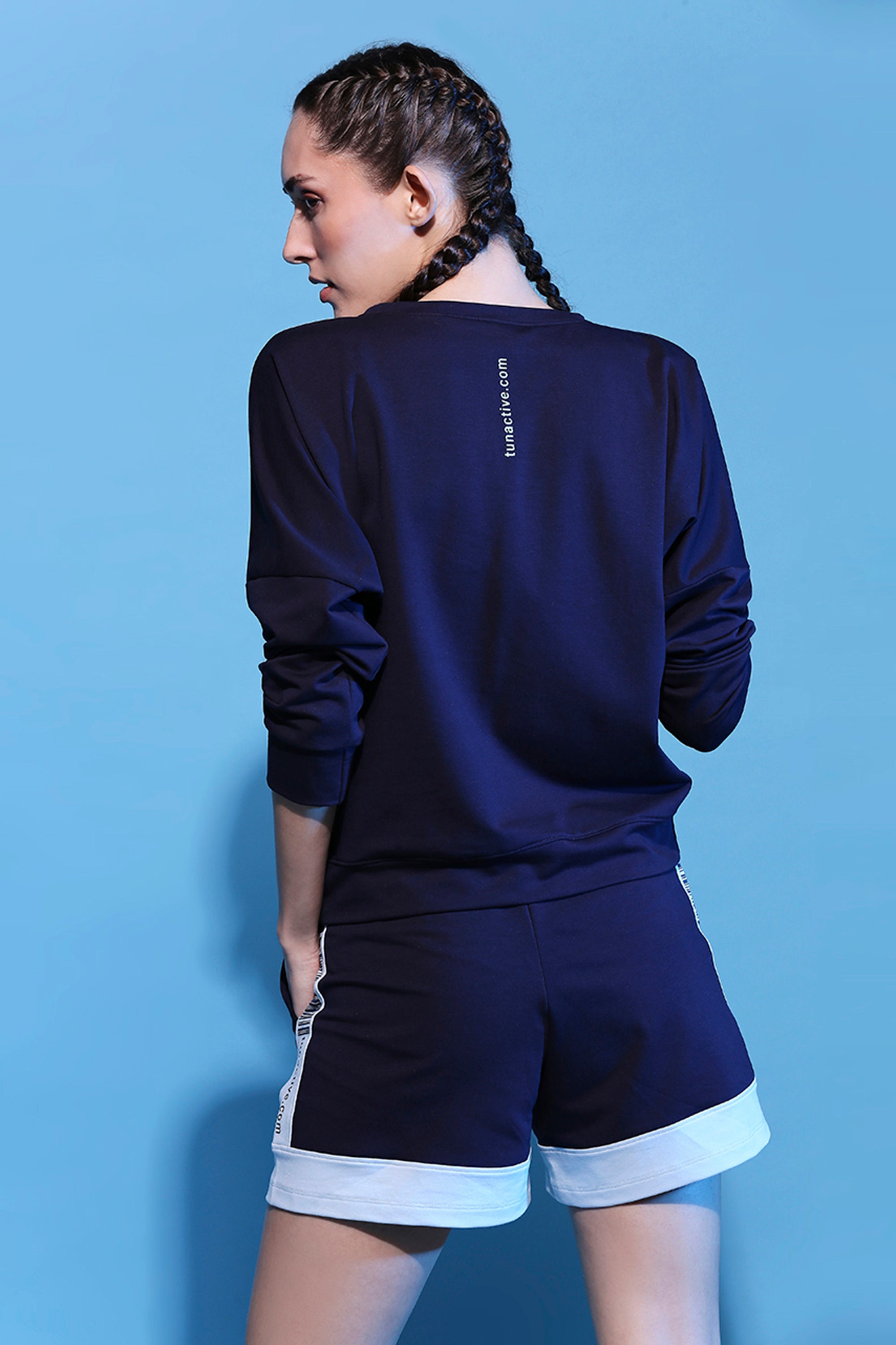 Woman with braided hair in the Barcode Printed Sweatshirt + Shorts - AW21 stands against a blue background, facing away—an effortless look from the Autumn Winter 21 Drop.