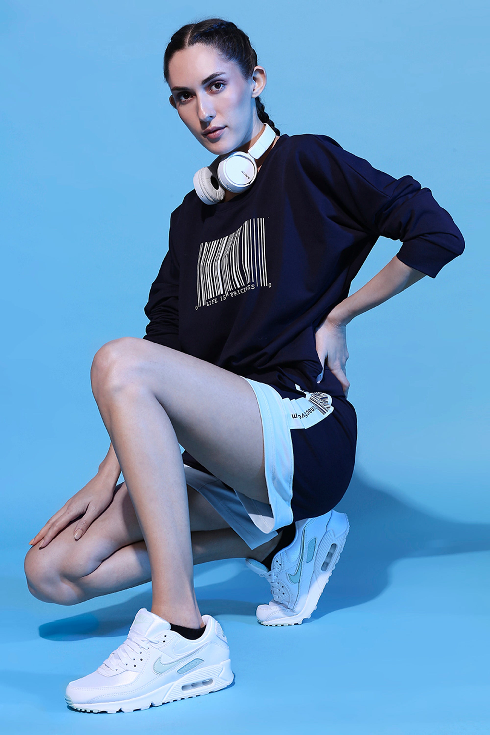 Woman in sporty Barcode Printed Sweatshirt + Shorts - AW21 outfit and headphones kneels on one knee against a blue background.