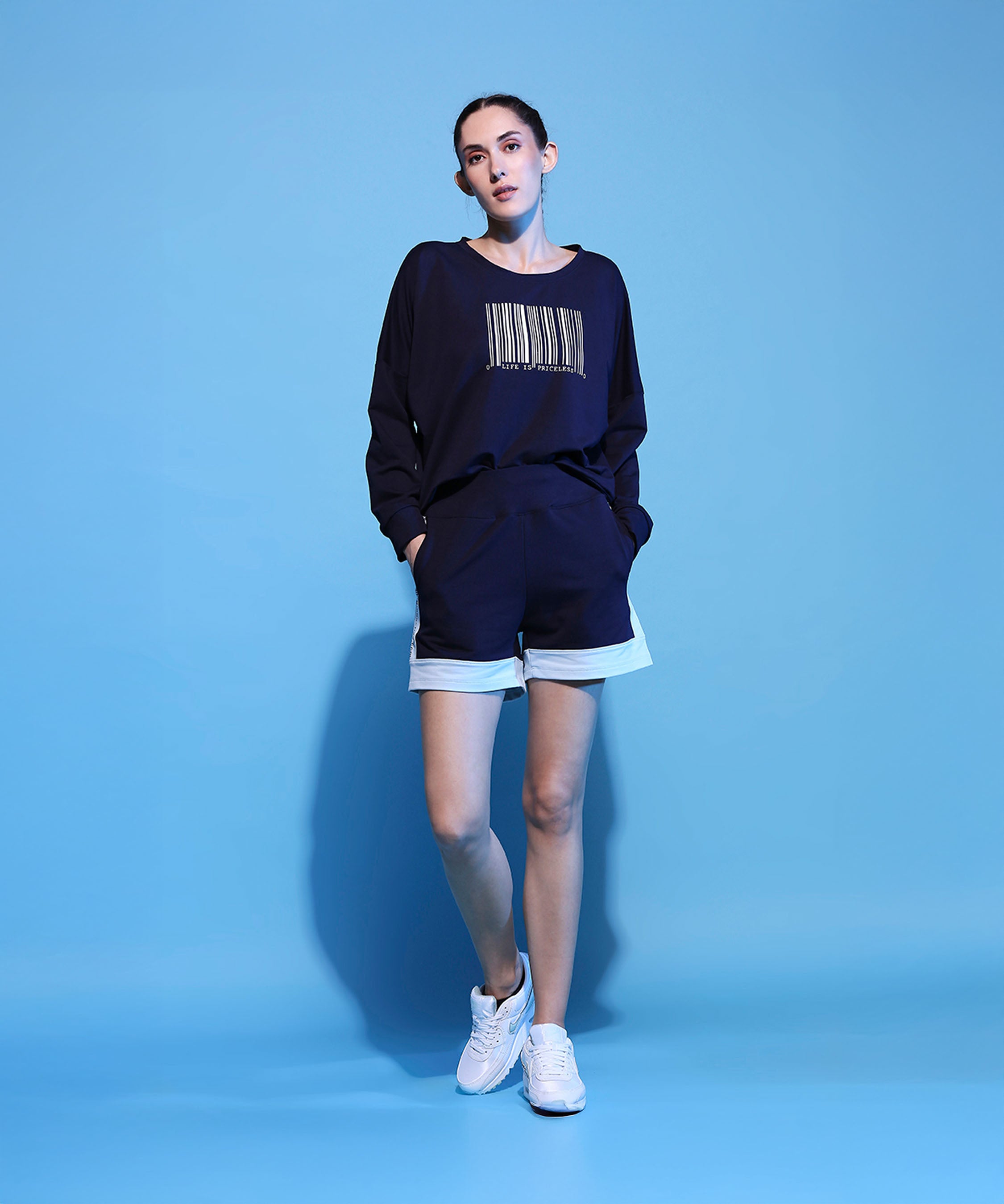 Woman in a Barcode Printed Navy Blue Sweatshirt and shorts, standing against a blue background with hands in pockets, wearing white sneakers.