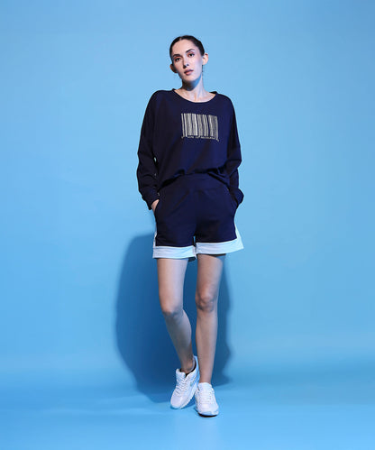 Woman in a Barcode Printed Navy Blue Sweatshirt and shorts, standing against a blue background with hands in pockets, wearing white sneakers.