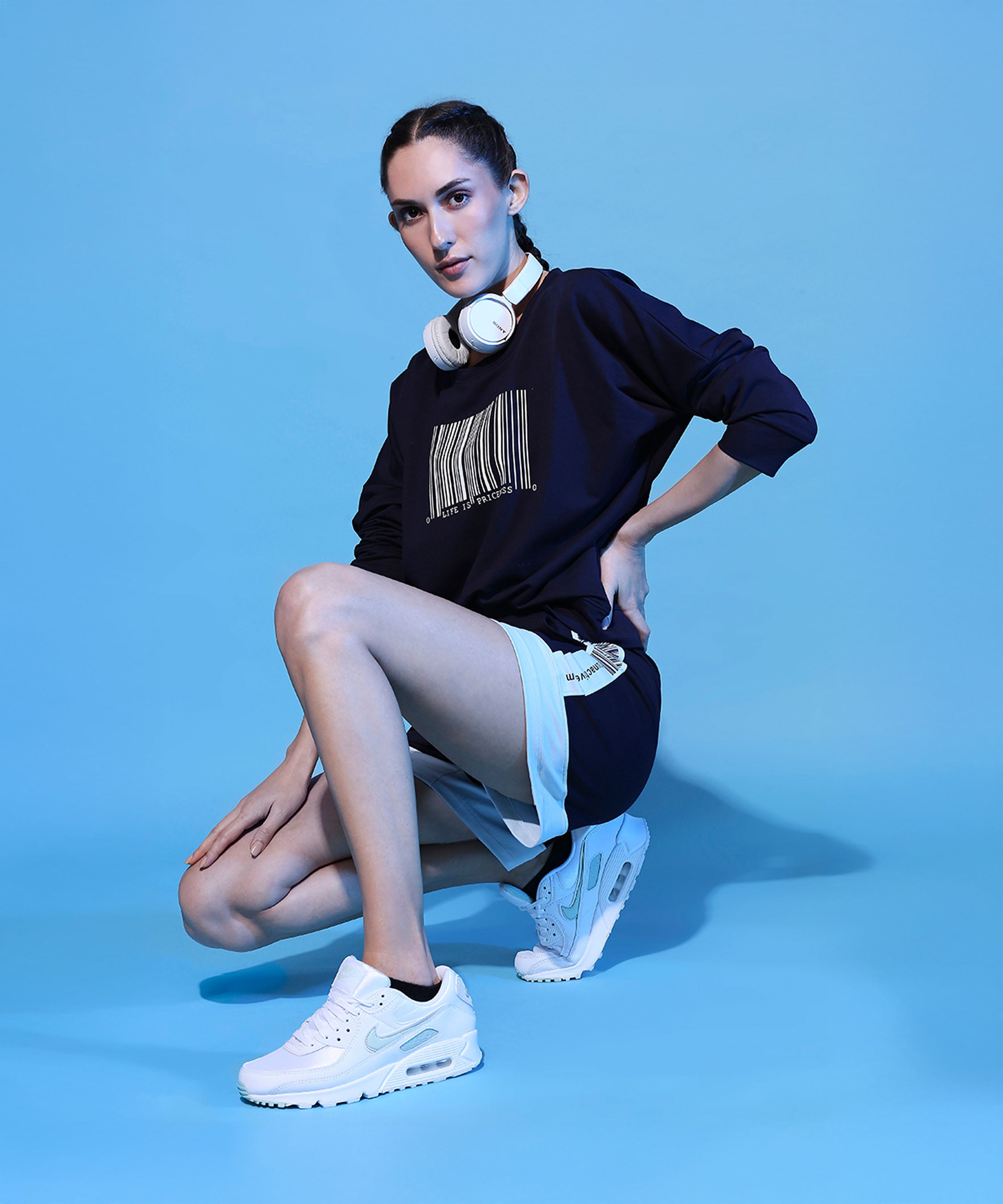 A woman in sporty attire, wearing a Barcode Printed Navy Blue Sweatshirt and white headphones, poses confidently against a blue background.