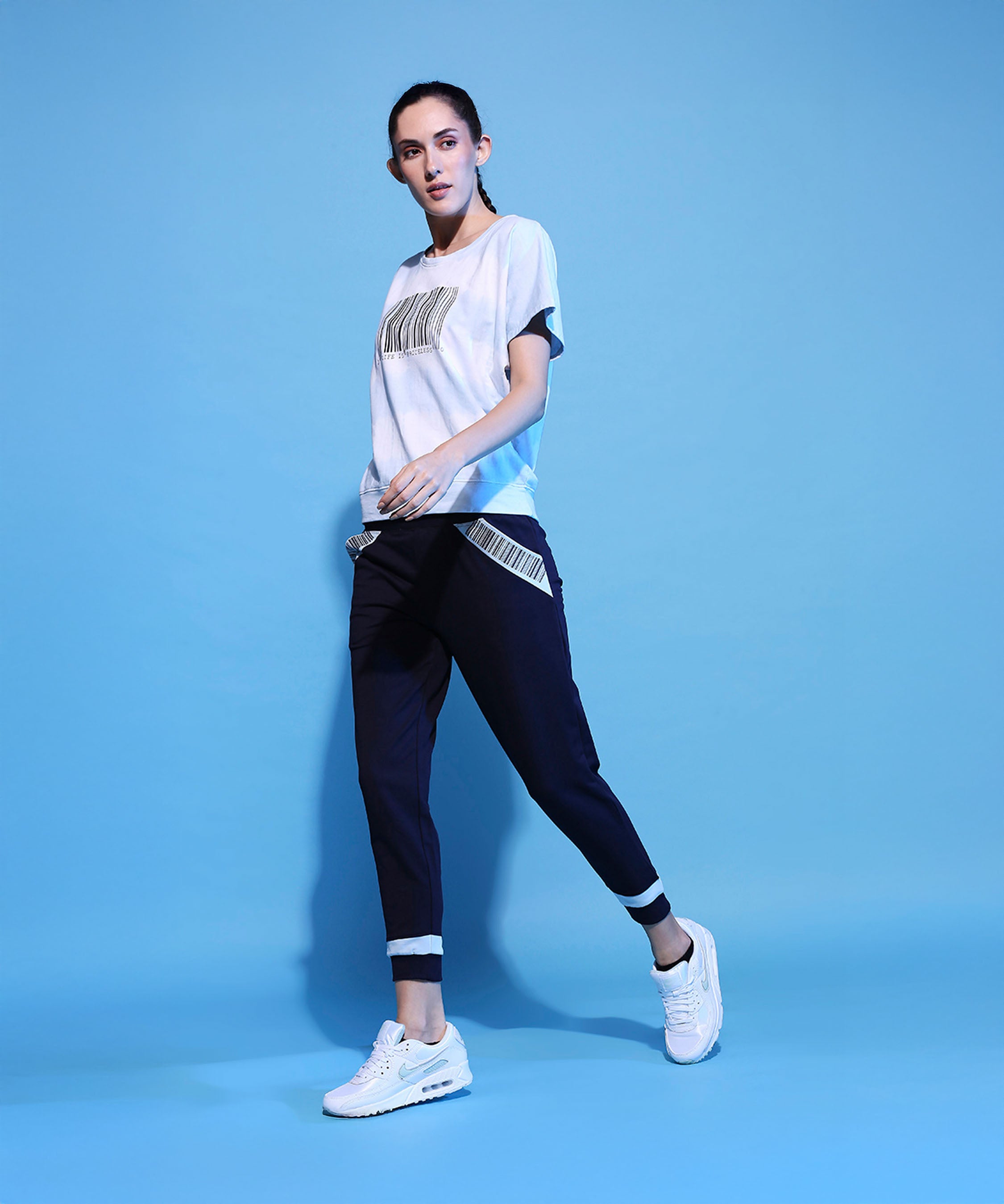 A woman models the Barcode Printed Comfort Tie Dye Tee + Joggers - AW21 with white sneakers, posing confidently against a solid light blue background.