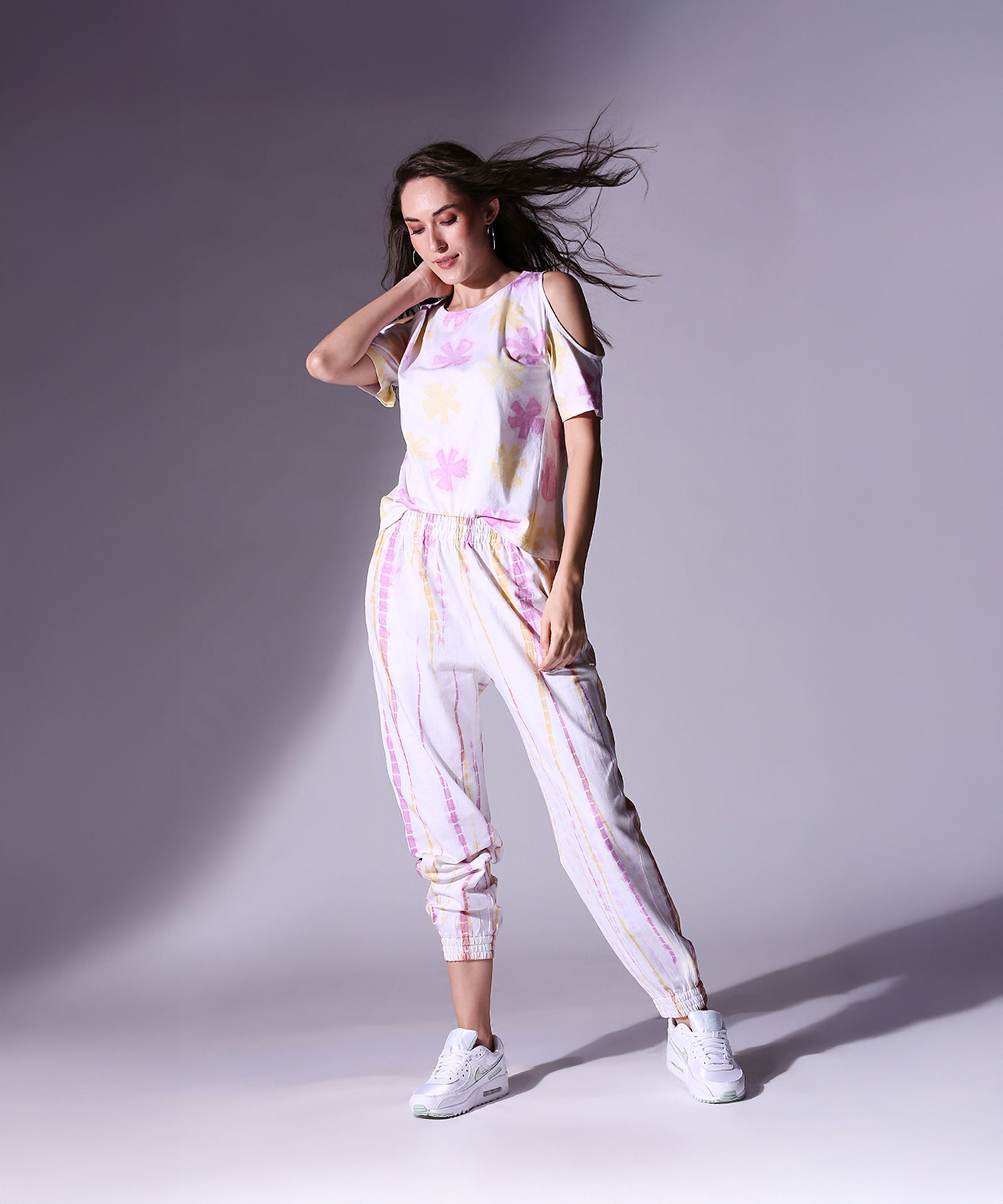 Woman modeling the AW21 Cold Shoulder Tee + Joggers set with white sneakers against a light gray studio background.