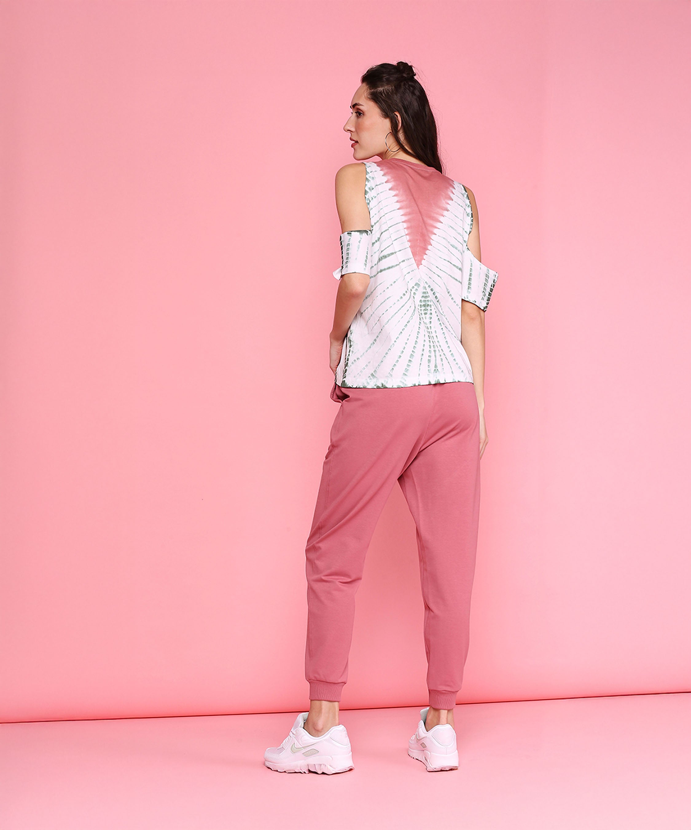 A woman stands against a pink background, facing away, wearing the Cold Shoulder Comfort Fit Top + Joggers - AW21 in carrot pink with white sneakers.