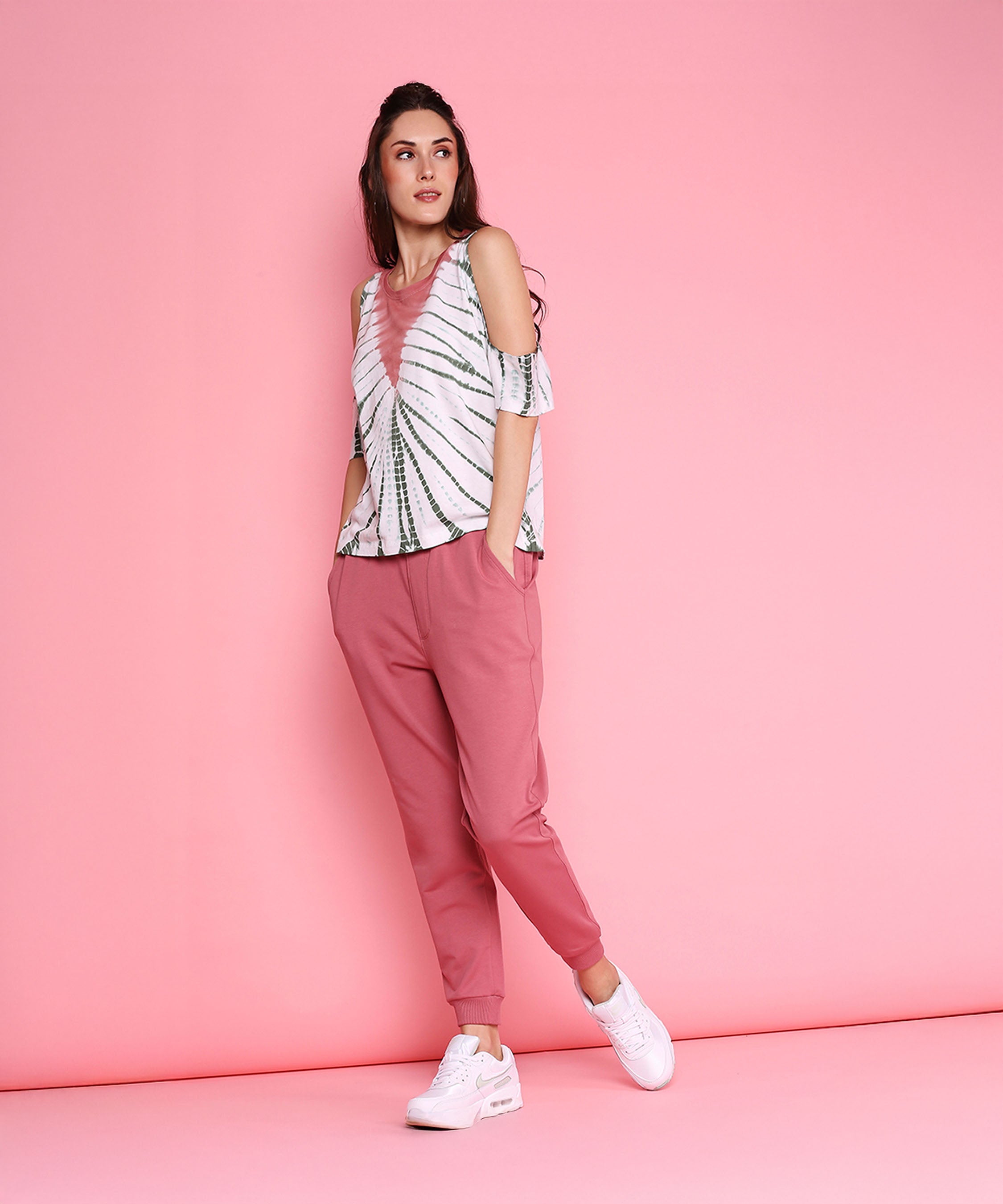 Woman in the Cold Shoulder Comfort Fit Top + Joggers - AW21, featuring a pink and white tie dye cold shoulder top, carrot pink joggers, and white sneakers, stands against a pink background.