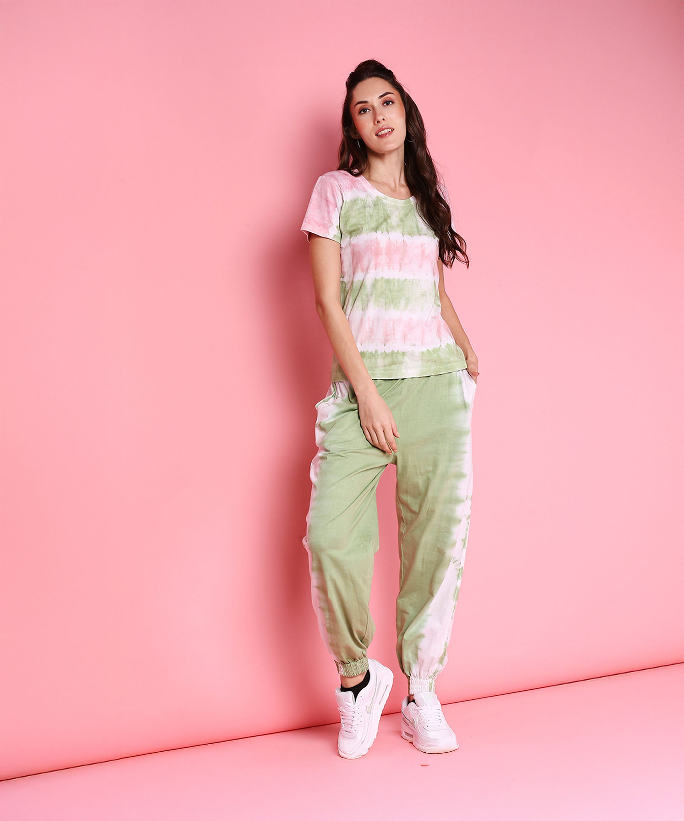Woman poses against a solid pink background in the Tee With Racer Back Cut + Joggers - AW21, styled with mint green joggers, a pastel pink tee, and white sneakers.