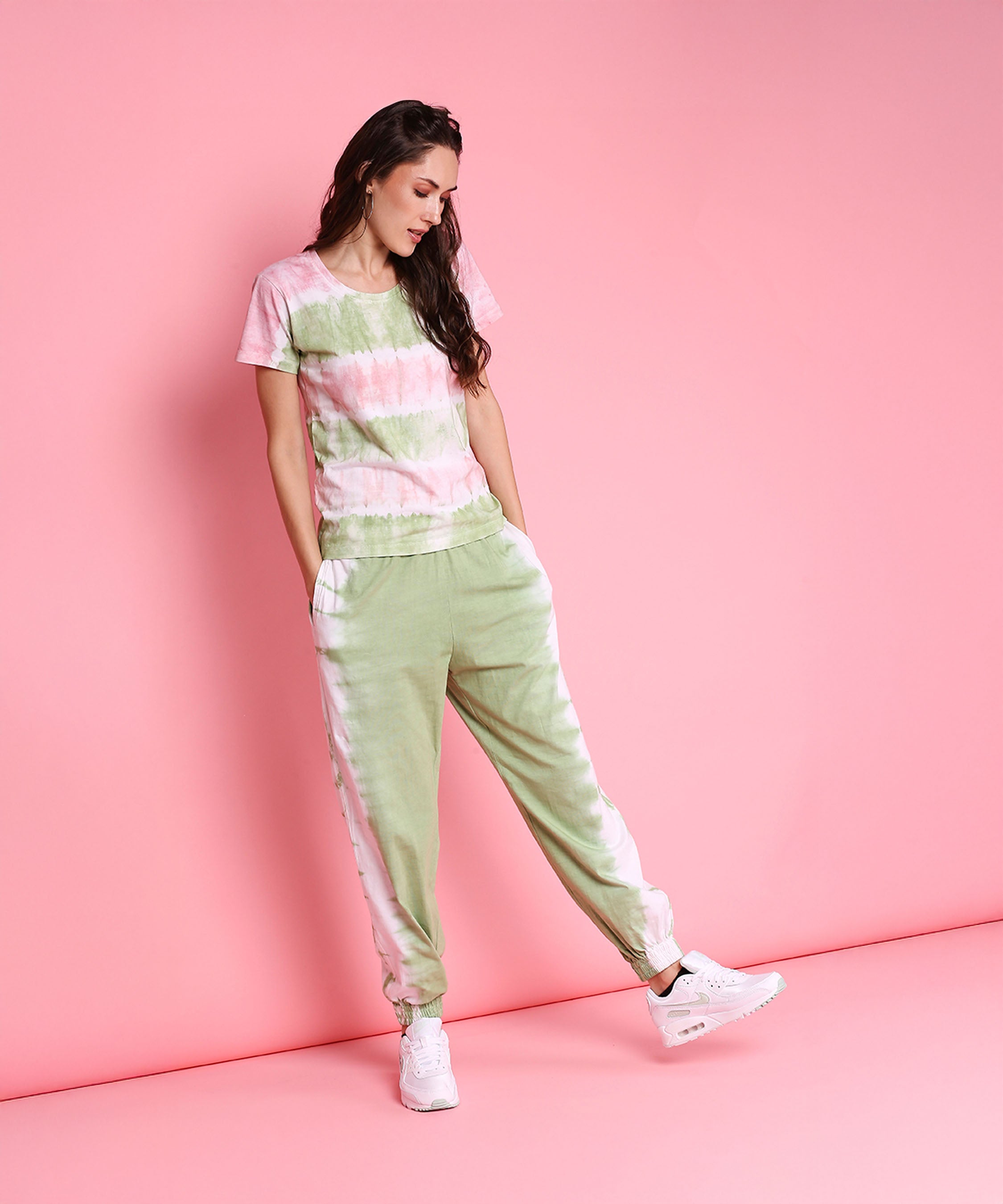A woman in the Tee With Racer Back Cut + Joggers - AW21, featuring mint green joggers and a pastel pink tee, stands against a pink background, looking down and wearing crisp white sneakers.