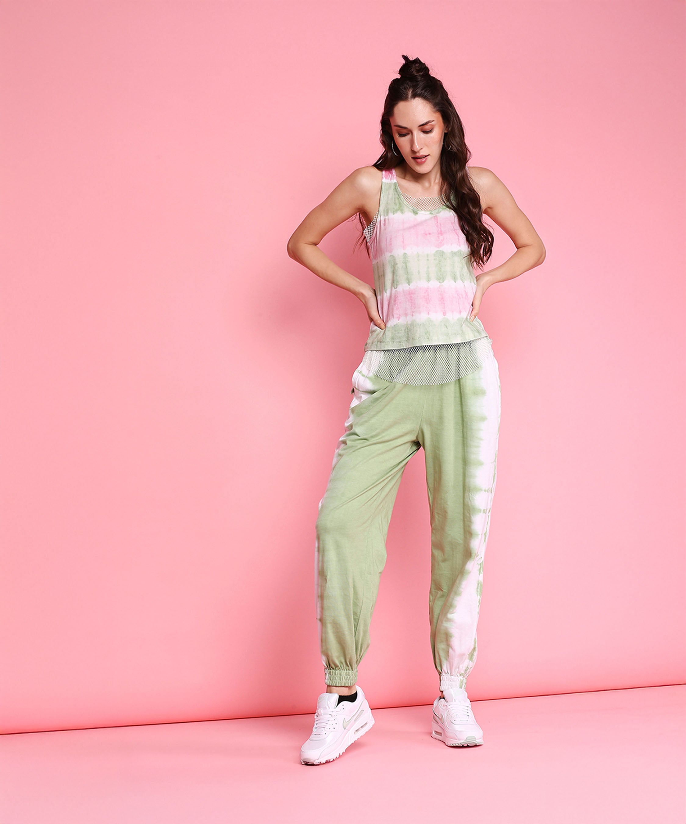 Woman models the Comfort Fit Joggers + Netted Tank Top - AW21, featuring a pink and green tie-dye design, against a solid pink background with white sneakers.