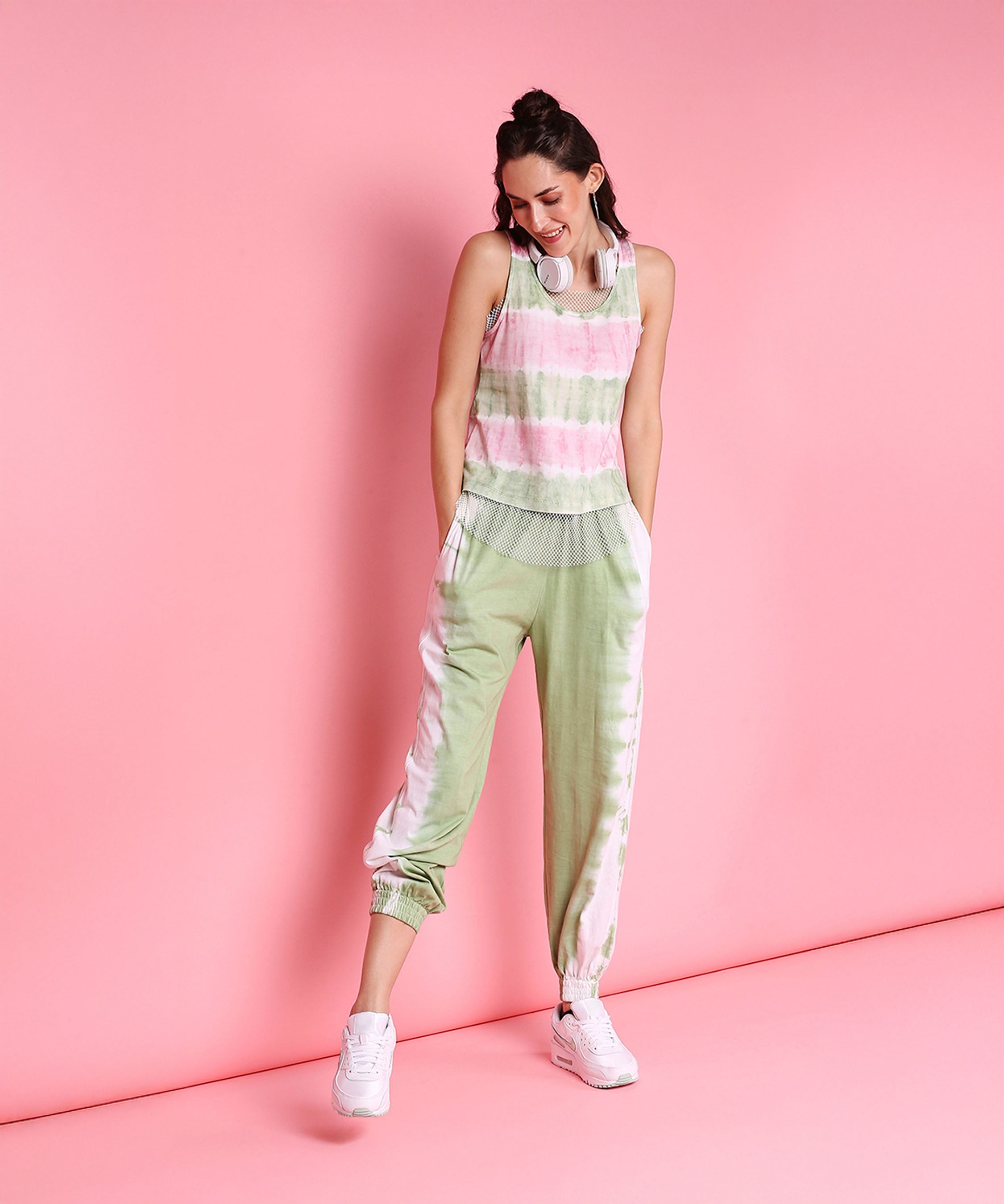Woman in the Comfort Fit Joggers + Netted Tank Top - AW21, featuring pink and mint green pastel hues, paired with white sneakers, stands smiling against a pink background.