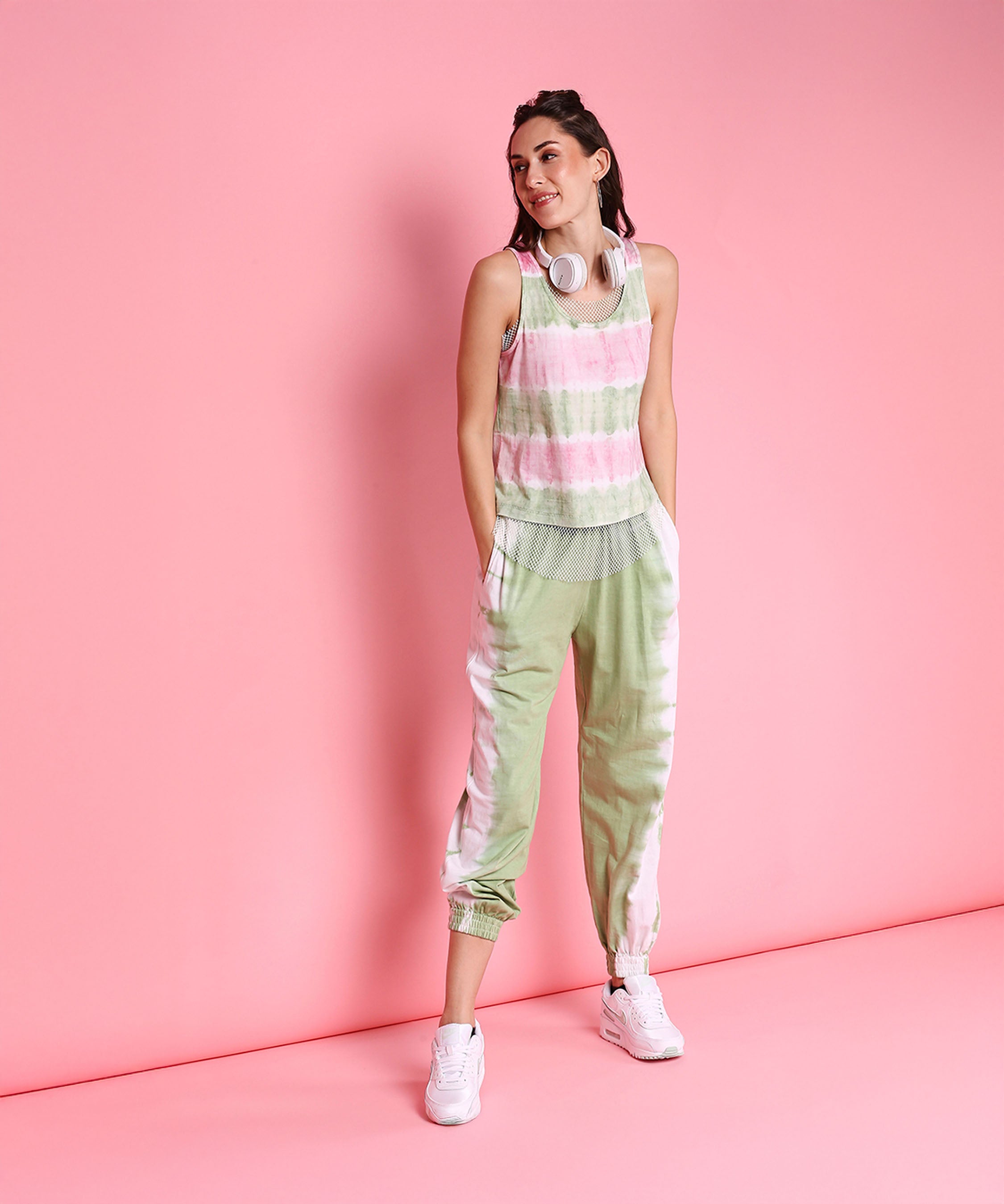 Woman wearing the Comfort Fit Joggers + Netted Tank Top - AW21 set with headphones around her neck, standing against a pink background.