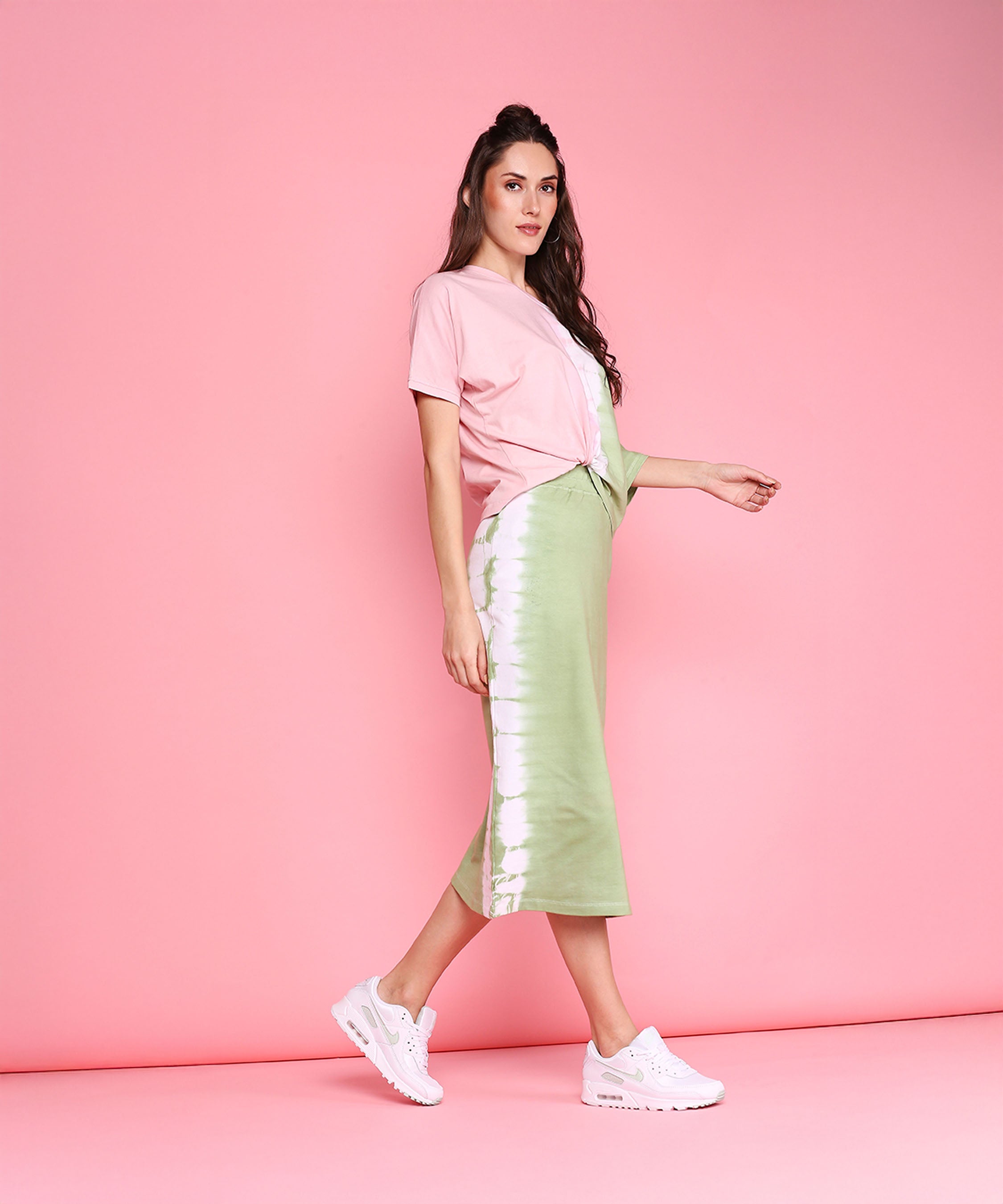 Woman in the Comfort Fit Tee and Fitted Tie Dye Skirt - AW21 poses against a pink background, completing her athleisure look with fresh white sneakers.