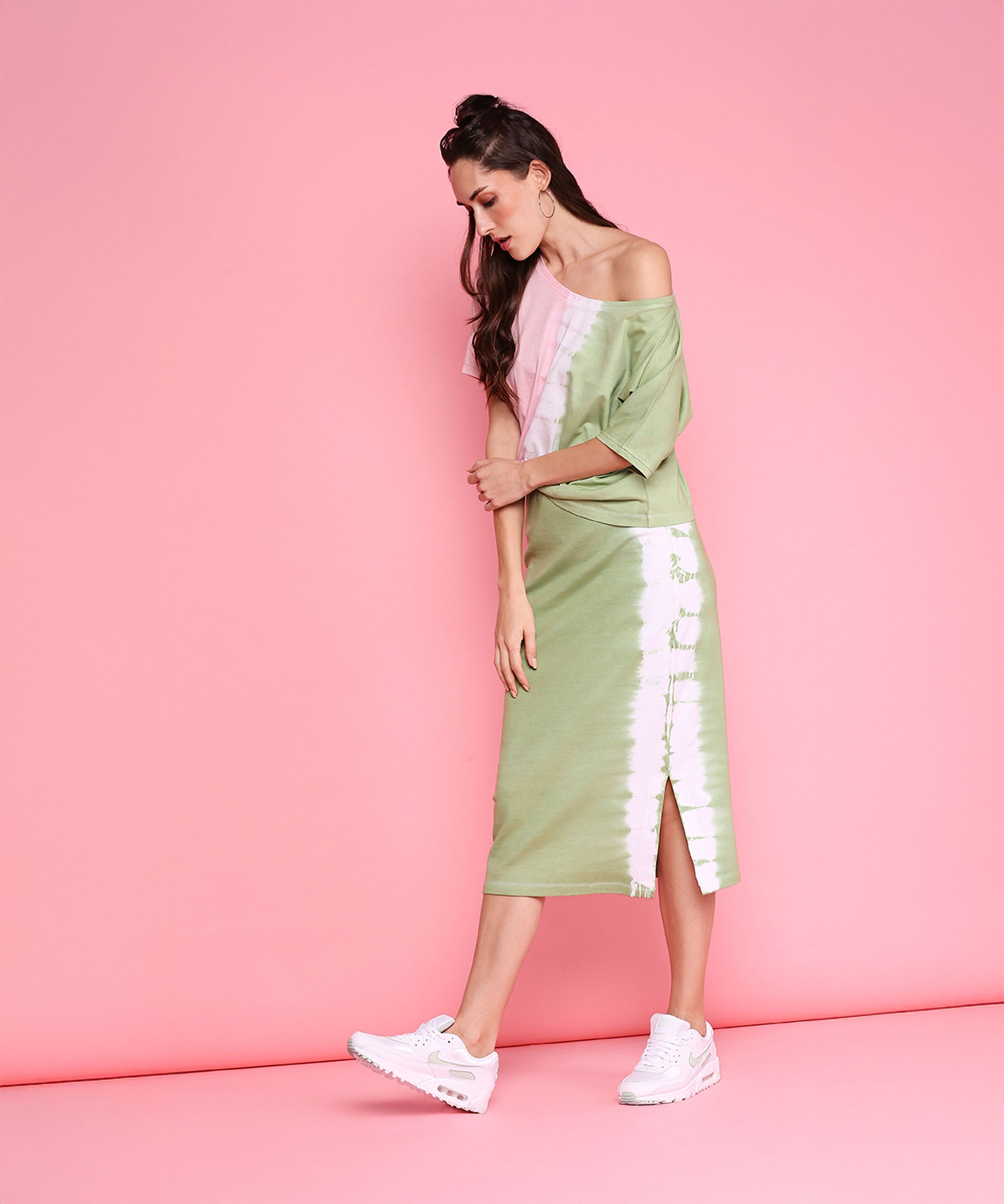 Woman wearing the Comfort Fit Tee and Fitted Tie Dye Skirt - AW21, featuring a pastel pink and mint green design with a side slit, styled with white sneakers in an athleisure look against a pink background.