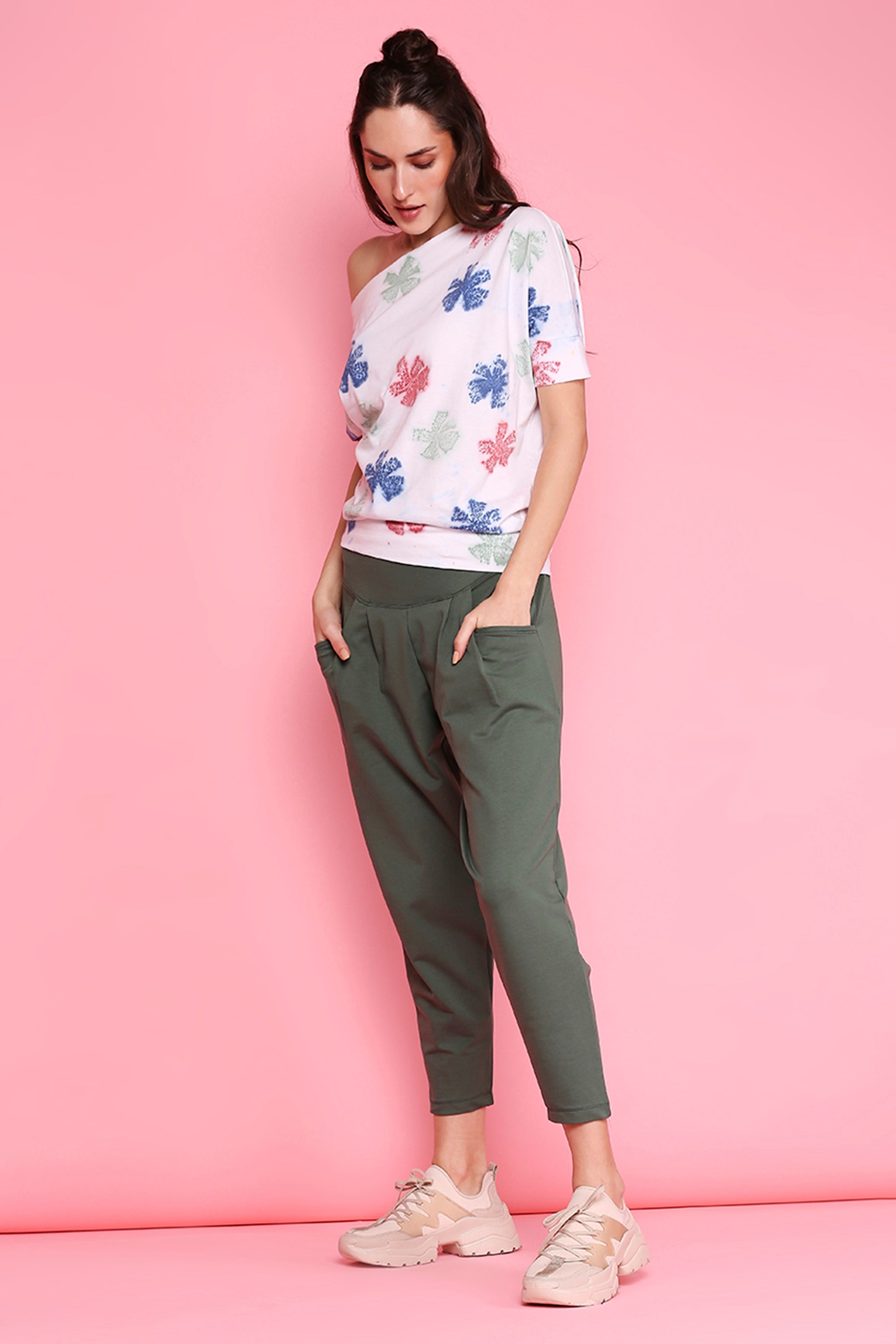 Woman in a floral top, Pleated Yoga Pants - AW21 (olive), and beige sneakers stands against a pink background with her hands in her pockets.