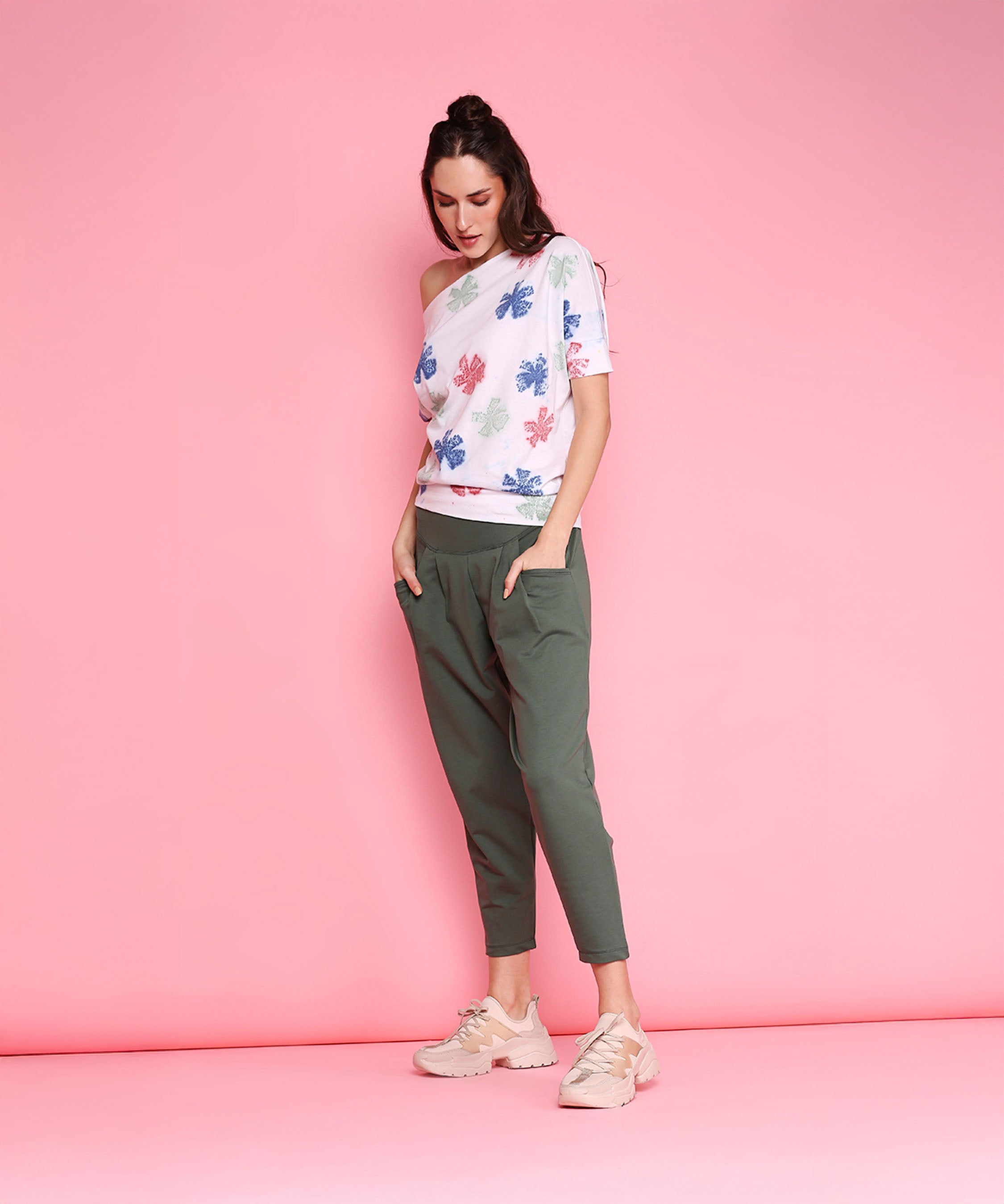 Woman wears the One Side Off Shoulder top and Pleated Yoga Pants - AW21 in olive, standing against a pink background, looking down with hands in pockets.
