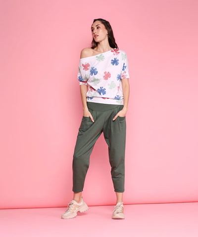 Woman in a floral off-shoulder top and Pleated Yoga Pants - AW21 with a green high waistband, standing against a pink background with hands in pockets.