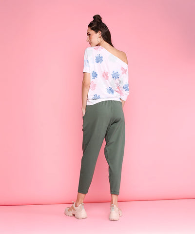 Woman faces away in Pleated Yoga Pants - AW21, featuring a high waistband and green harem style, paired with an off-shoulder floral top and sneakers, standing against a pink background.