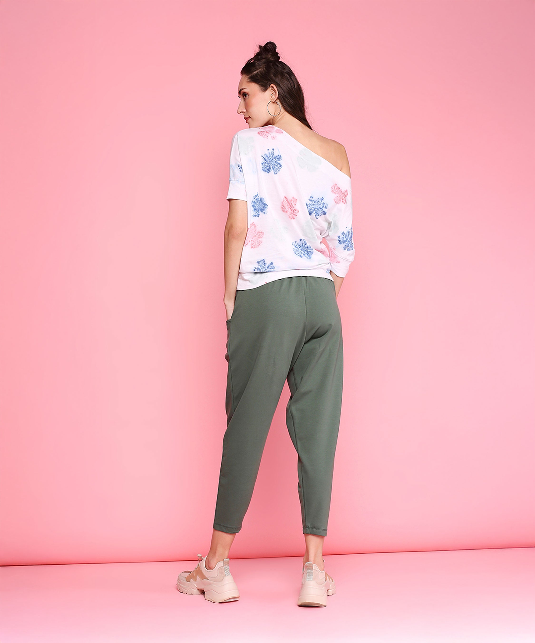 Woman stands against a pink background, wearing the One Side Off Shoulder + Pleated Yoga Pants - AW21 set in olive, paired with beige sneakers, facing away.