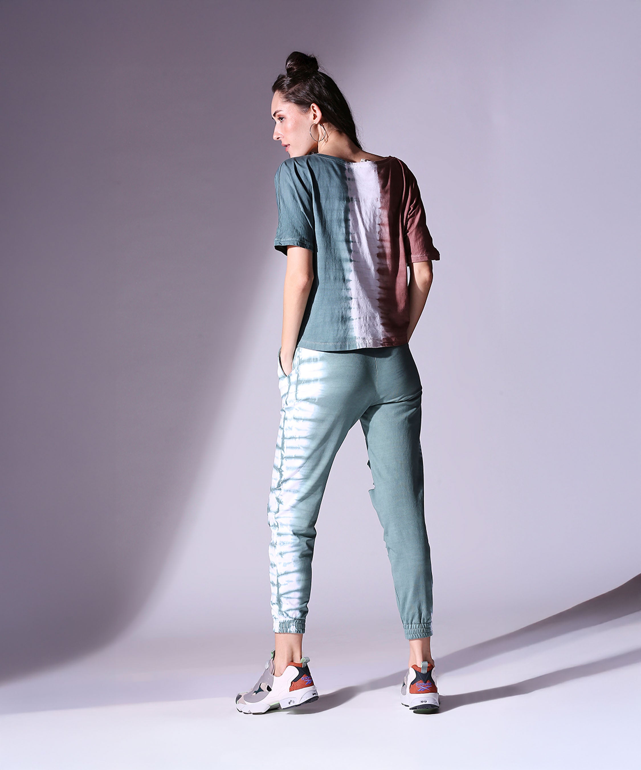 A woman models the Comfort Fit Tie Dye Top and Ripped Tie Dye Joggers - AW21 with sneakers, posing with her back turned in a softly lit studio for a cozy autumn-winter look.