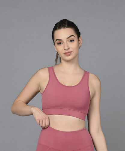 A woman in a Peony Pink Sports Bra and matching leggings stands against a gray background, holding the waistband of her moisture-wicking leggings.