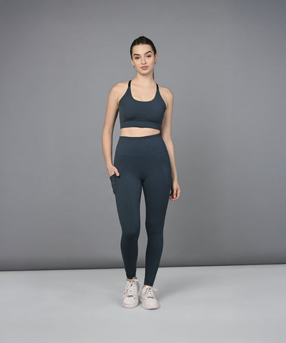A woman models the Teal Netted Top with blue leggings, standing with one hand in her pocket against a gray background.
