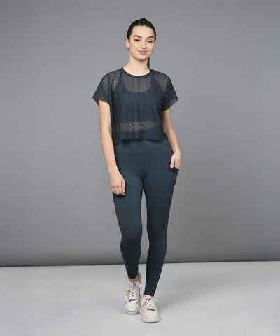 Woman wearing a Teal Netted Top with high-waisted leggings and sneakers, standing against a plain gray background.