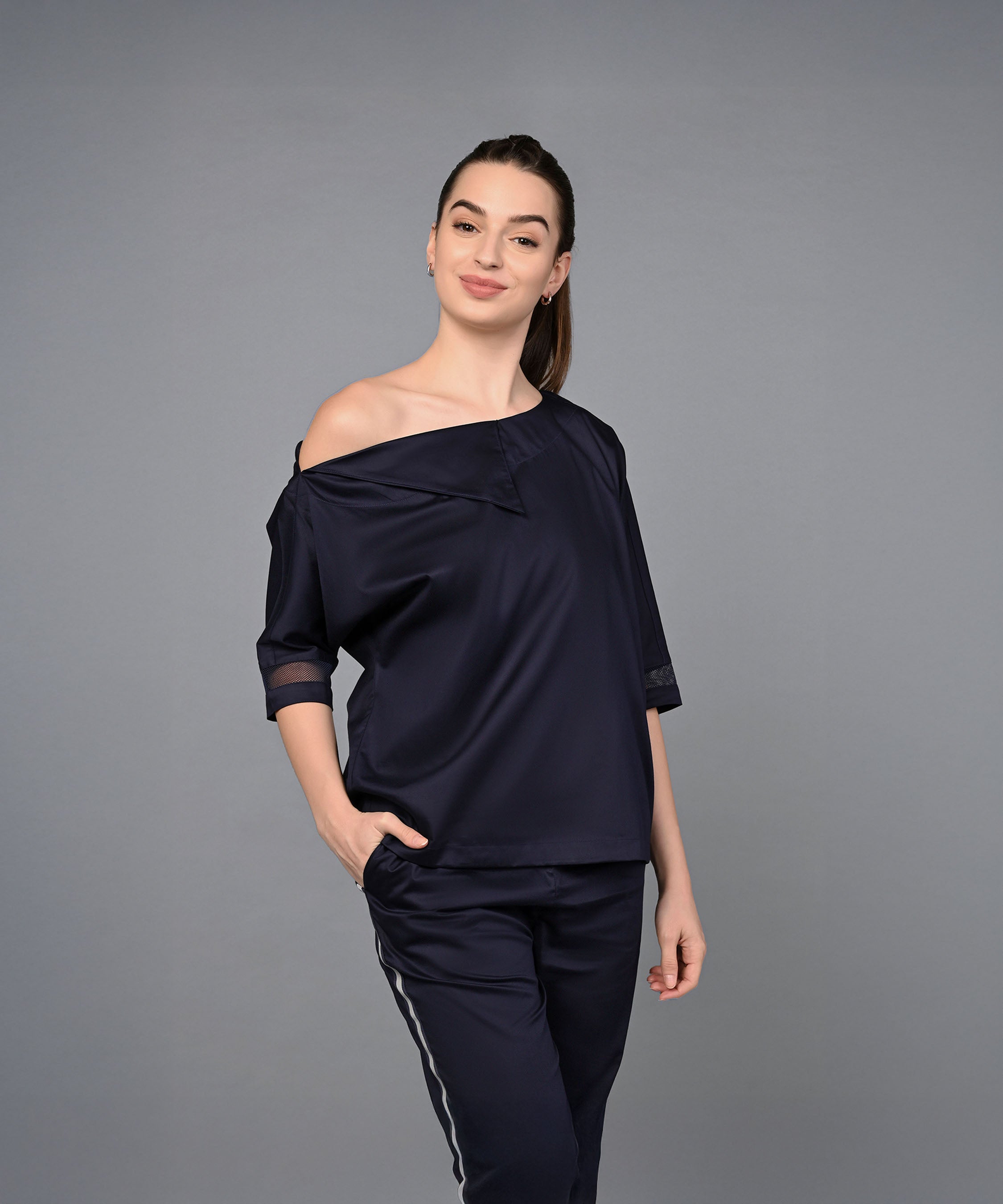 A woman wears the Off Shoulder Nebula Navy Co-ord set made from 100% cotton, standing with one hand in her pocket and smiling against a plain gray background.