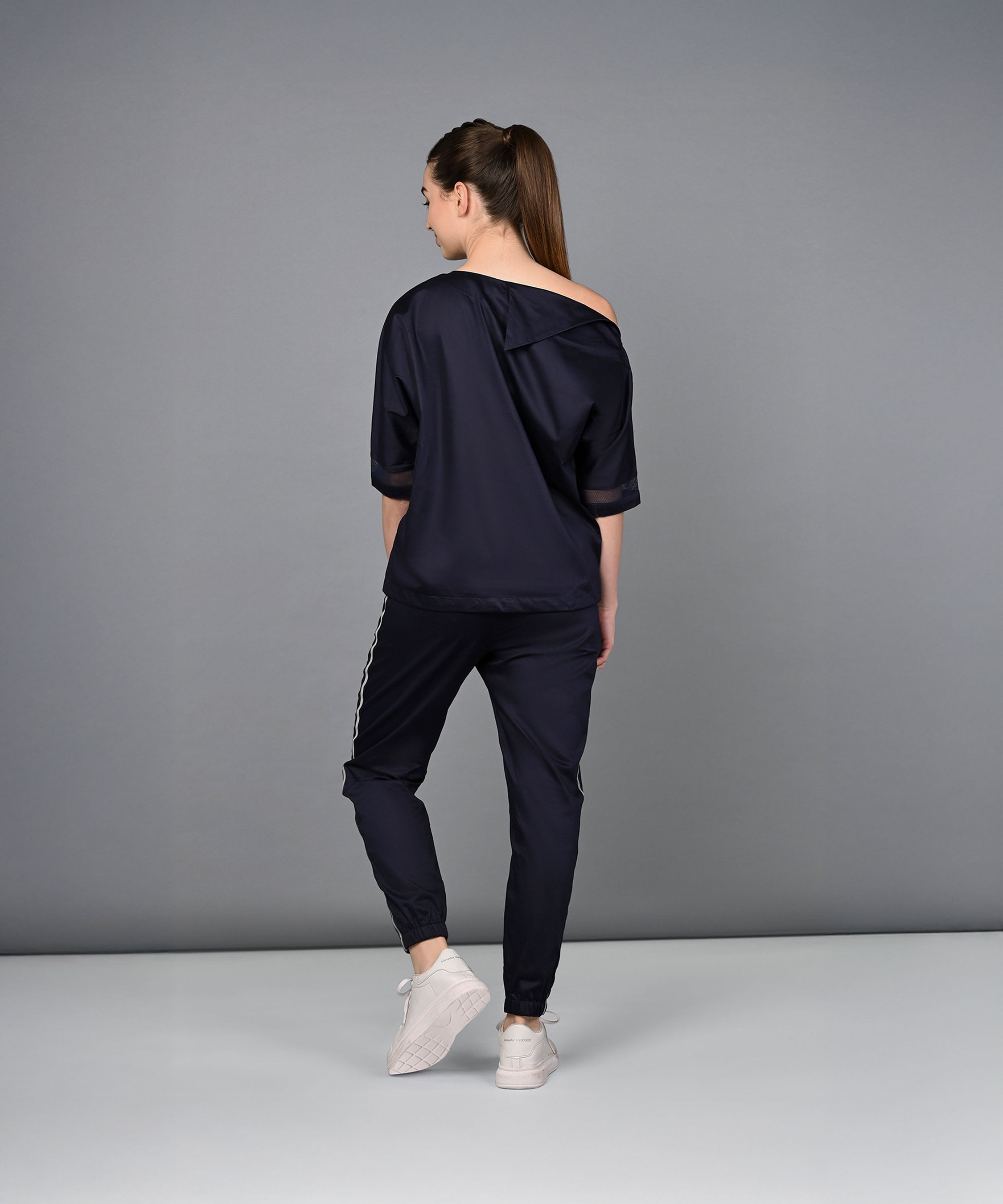 A woman wearing the Off Shoulder Nebula Navy Co-ord set, crafted from 100% cotton and paired with white sneakers, stands with her back to the camera against a gray background.