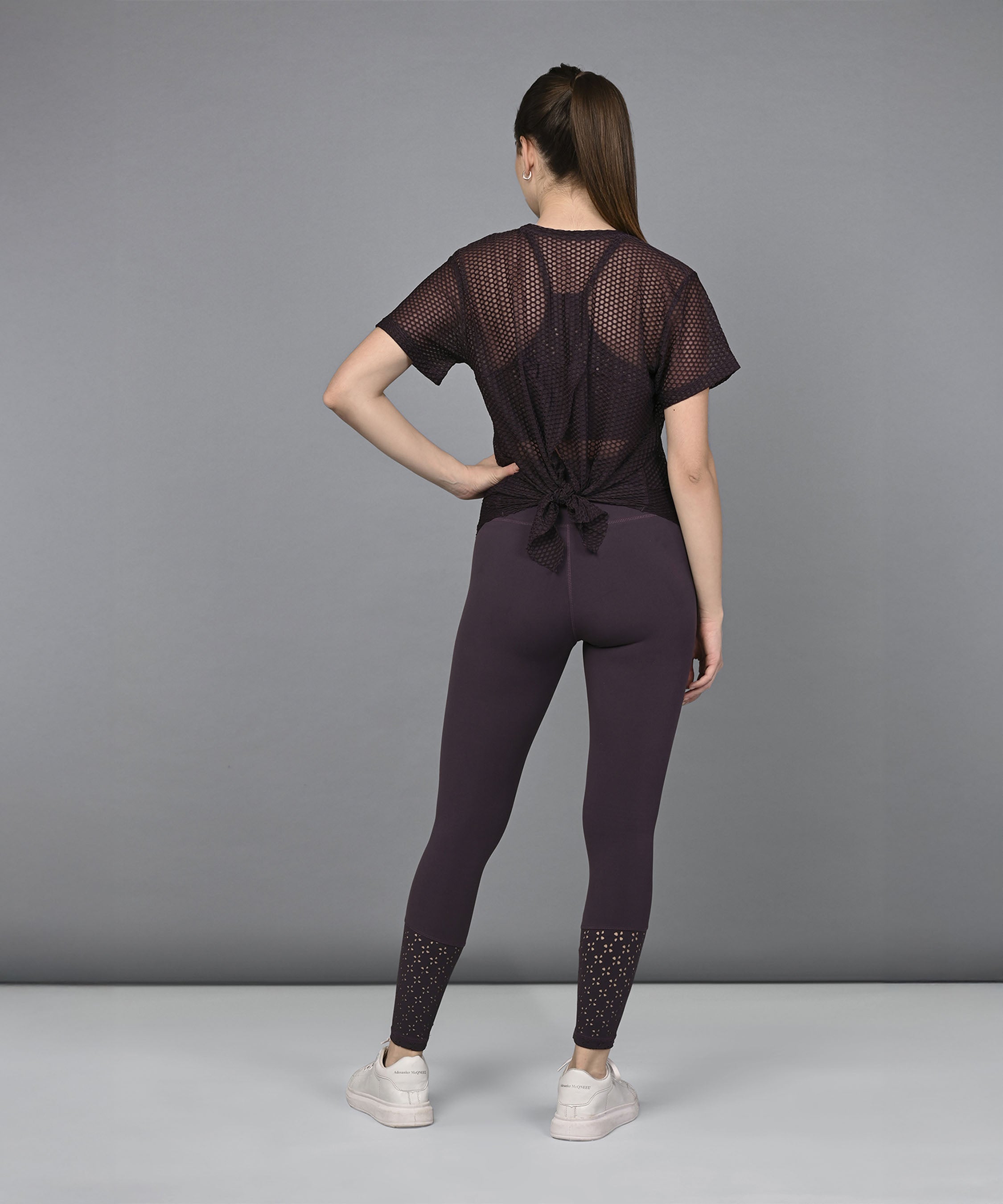 Woman in activewear stands facing away, wearing a sexy Wine Netted Top, purple leggings, and white sneakers.